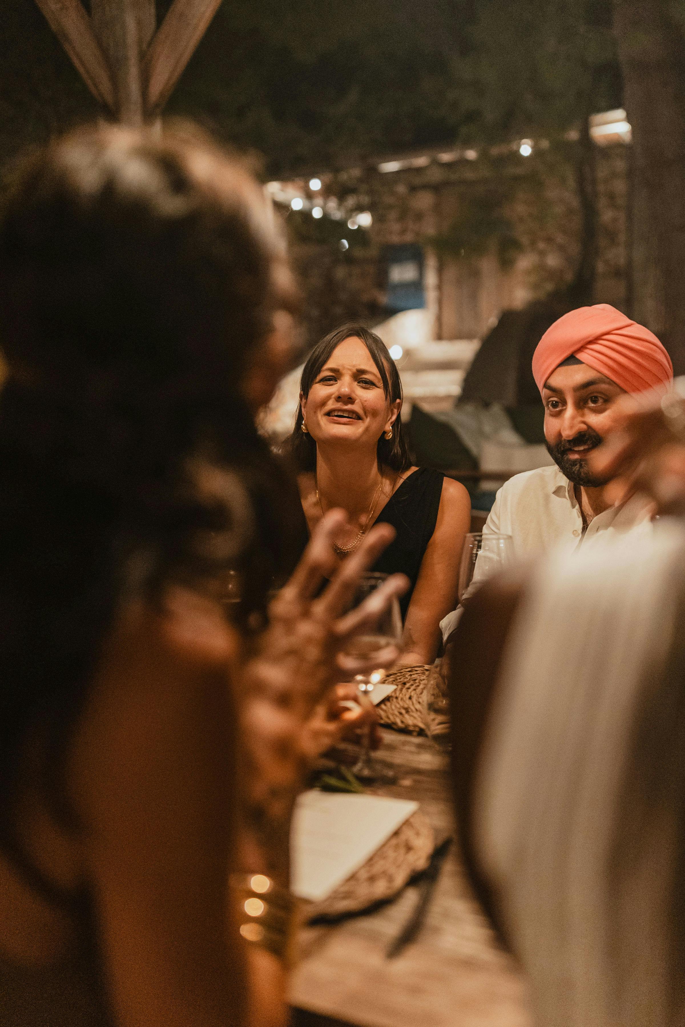 Wedding guests smiling and enjoying dinner at an outdoor evening reception. A woman in black and a person in white with a coral pink turban are visible in focus, while other guests are softly blurred in the foreground. The warm, intimate atmosphere is lit by candlelight and string lights overhead.