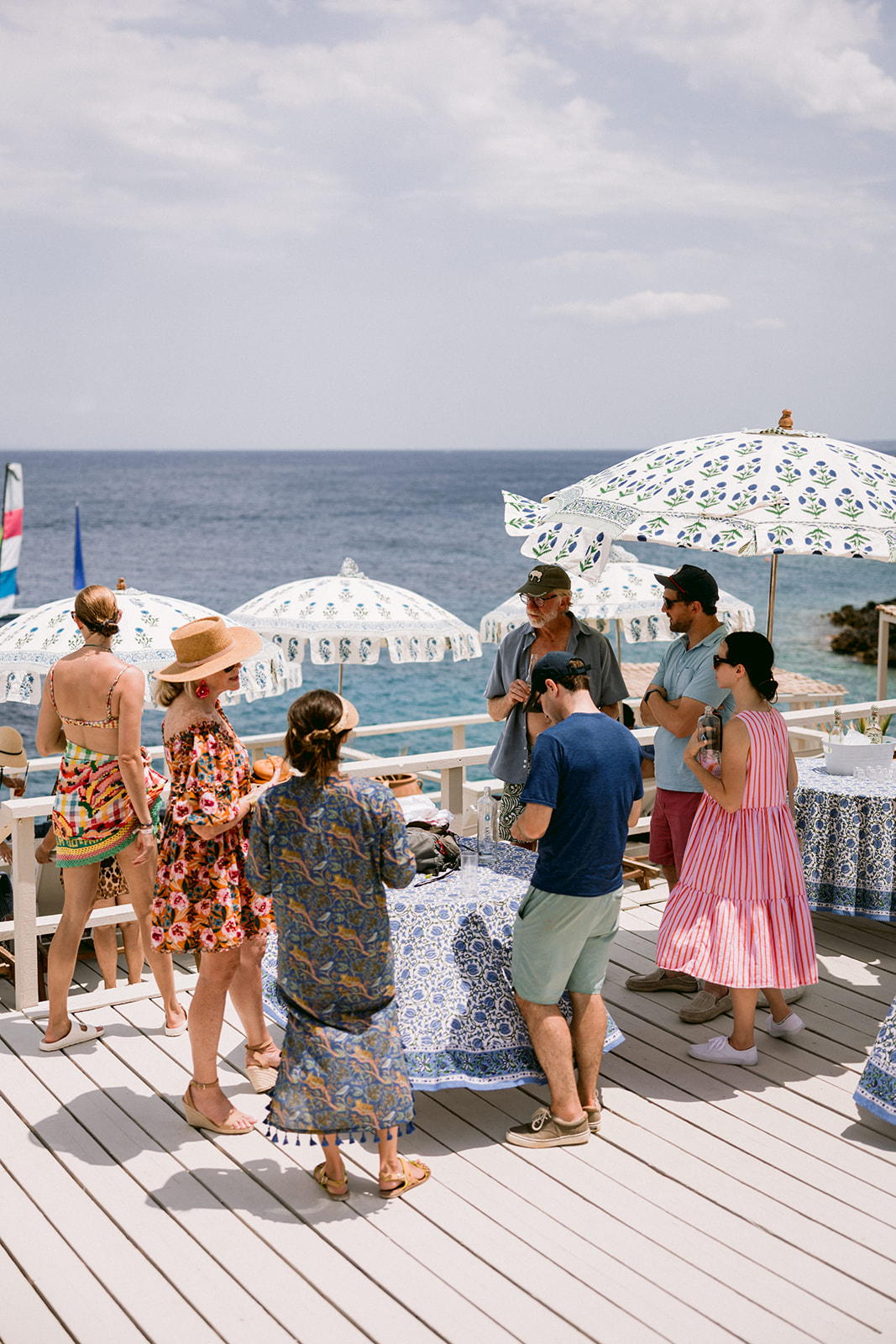 Wedding guests dancing on a white wooden deck overlooking the Mediterranean Sea. Guests in colourful summer attire dance together under decorative patterned umbrellas, with white lounge chairs visible in the background. The casual waterfront celebration takes place on a sunny day with ocean horizon visible under cloudy skies.
