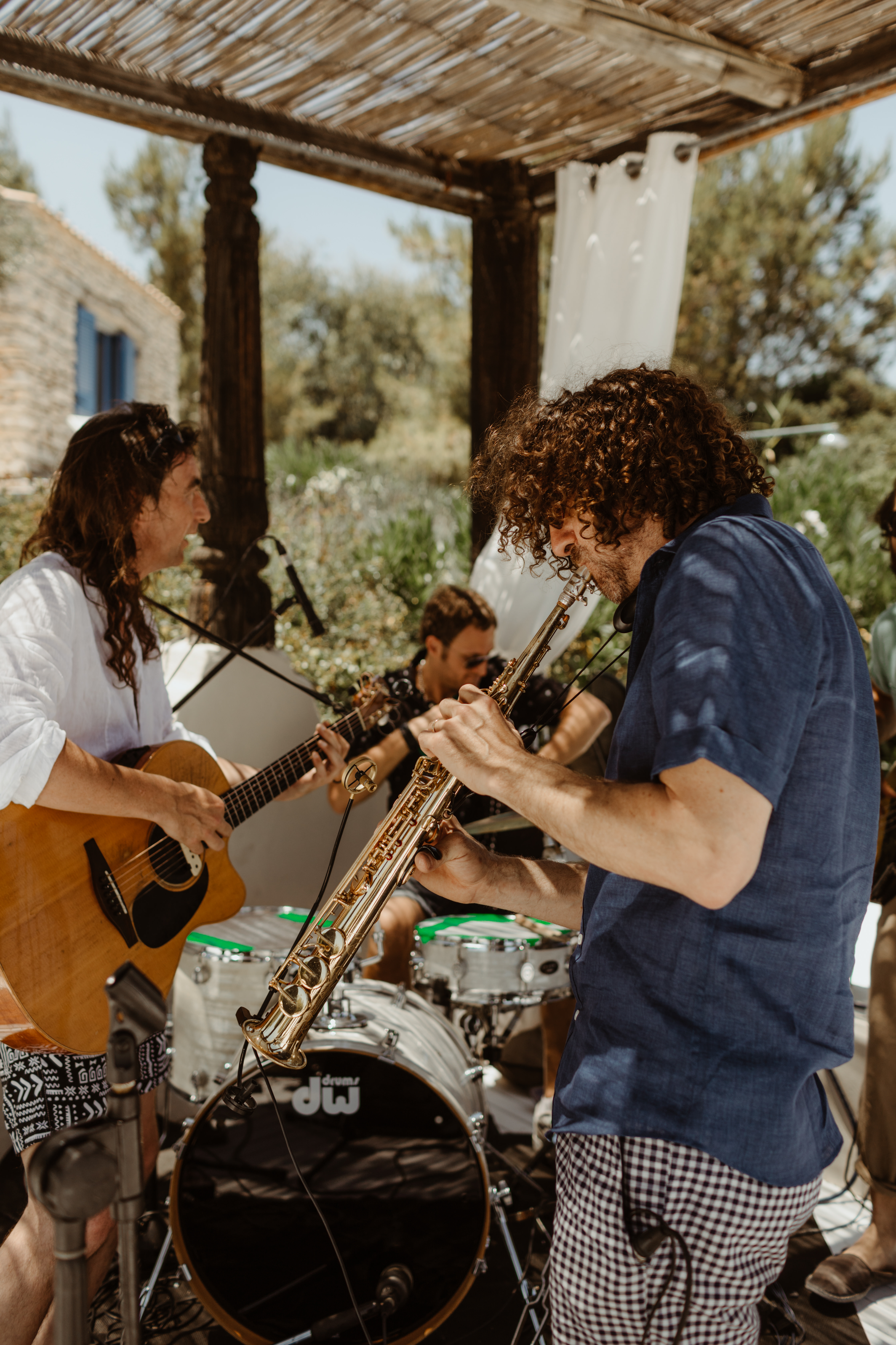 Live musicians performing at outdoor wedding reception under bamboo pergola. The guitarist is in a white shirt and saxophonist is in blue patterned shirt. They play together, with drum kit visible between them. The intimate performance takes place in a natural garden setting with olive trees in the background.