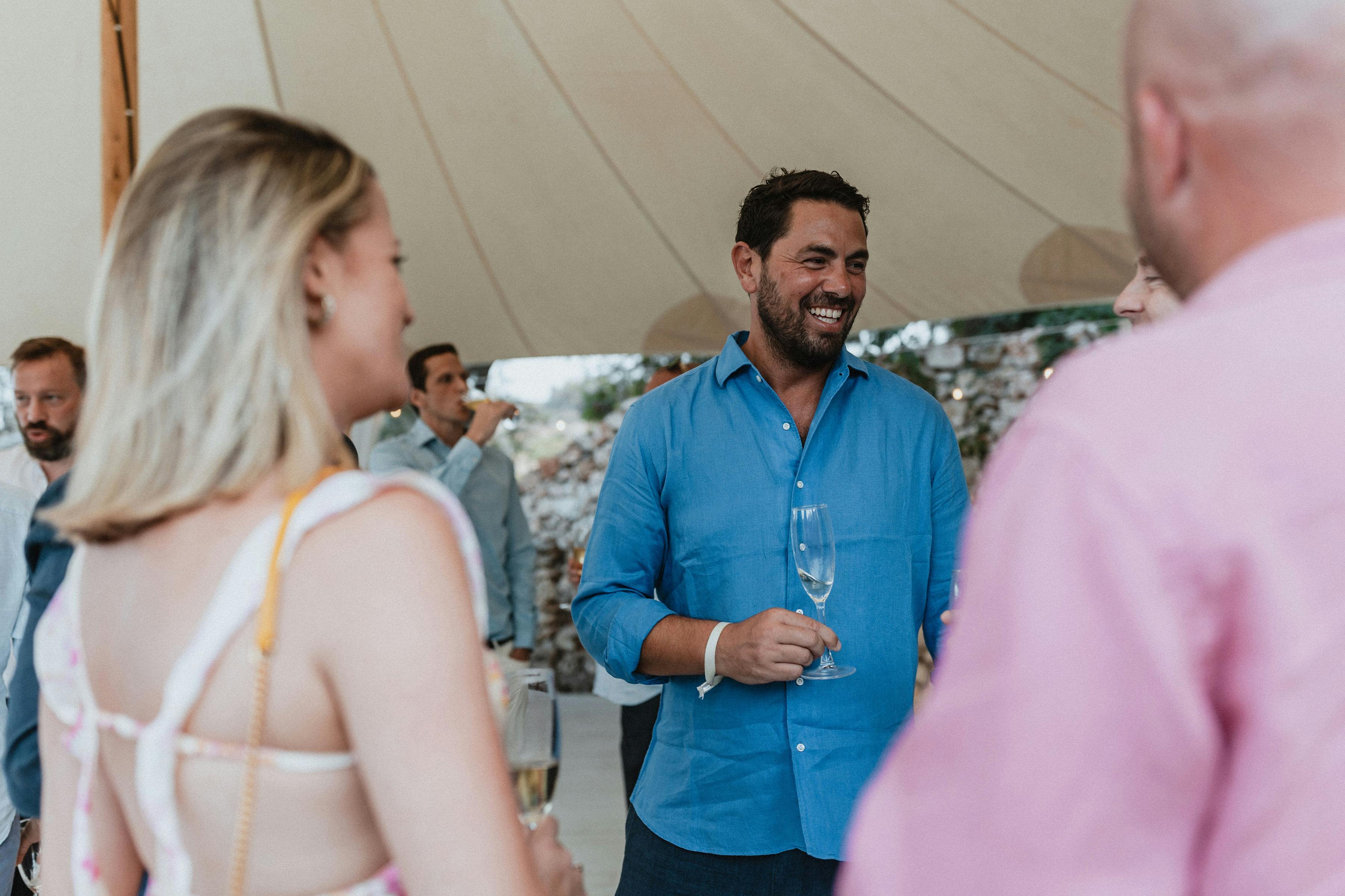 Guest in bright blue shirt smiling while holding wine glass and conversing with other guests at an outdoor reception under a white tent. The relaxed atmosphere captures guests mingling during a casual celebration, with stone walls visible in the background.