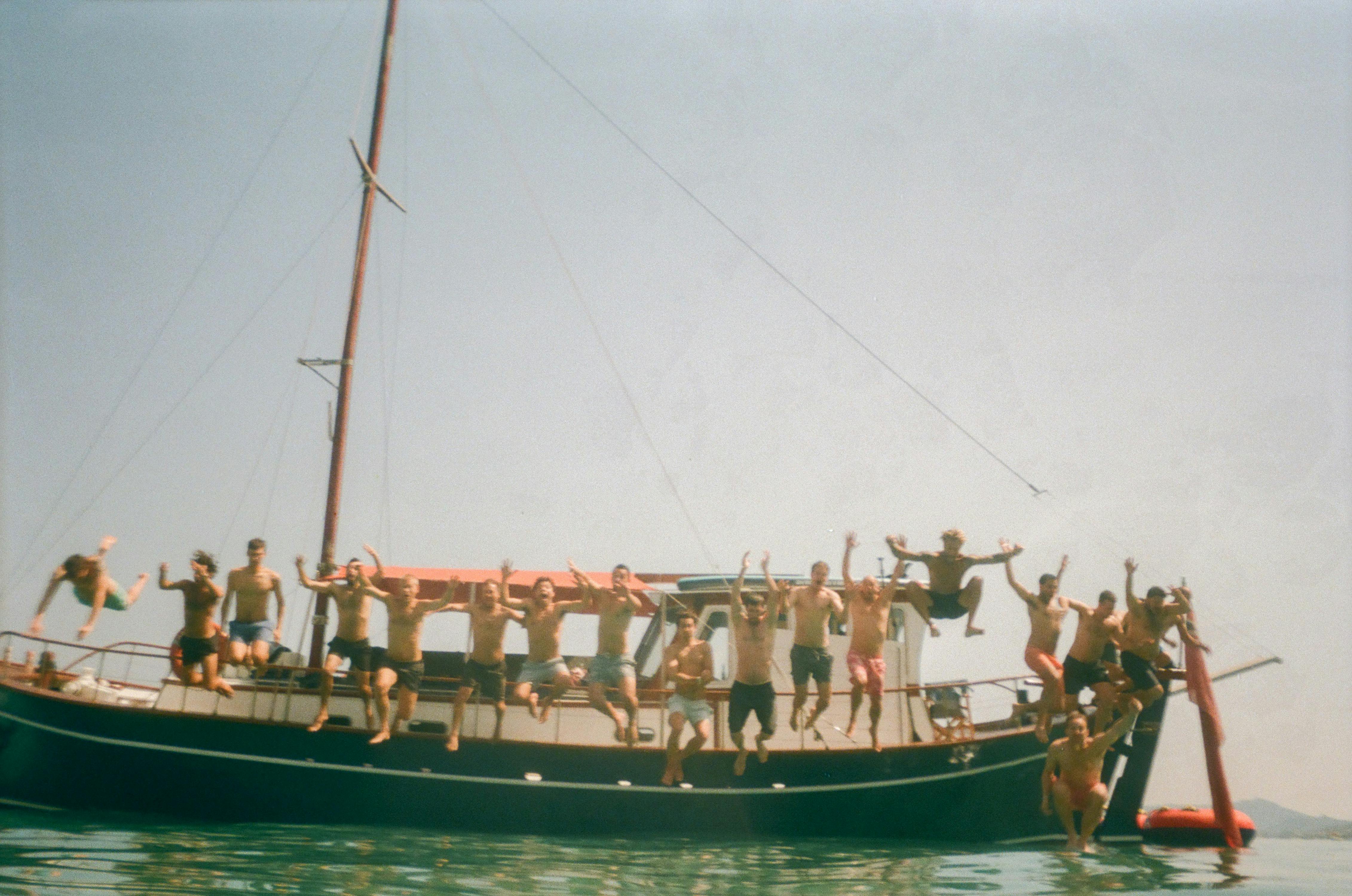 Guests in swimwear crowded on the deck of a large wooden sailboat in calm waters. Multiple people stand along the boat's edge and on deck with arms raised in celebration, enjoying a group boat excursion during a destination wedding. The mast and rigging are visible against a hazy sky.