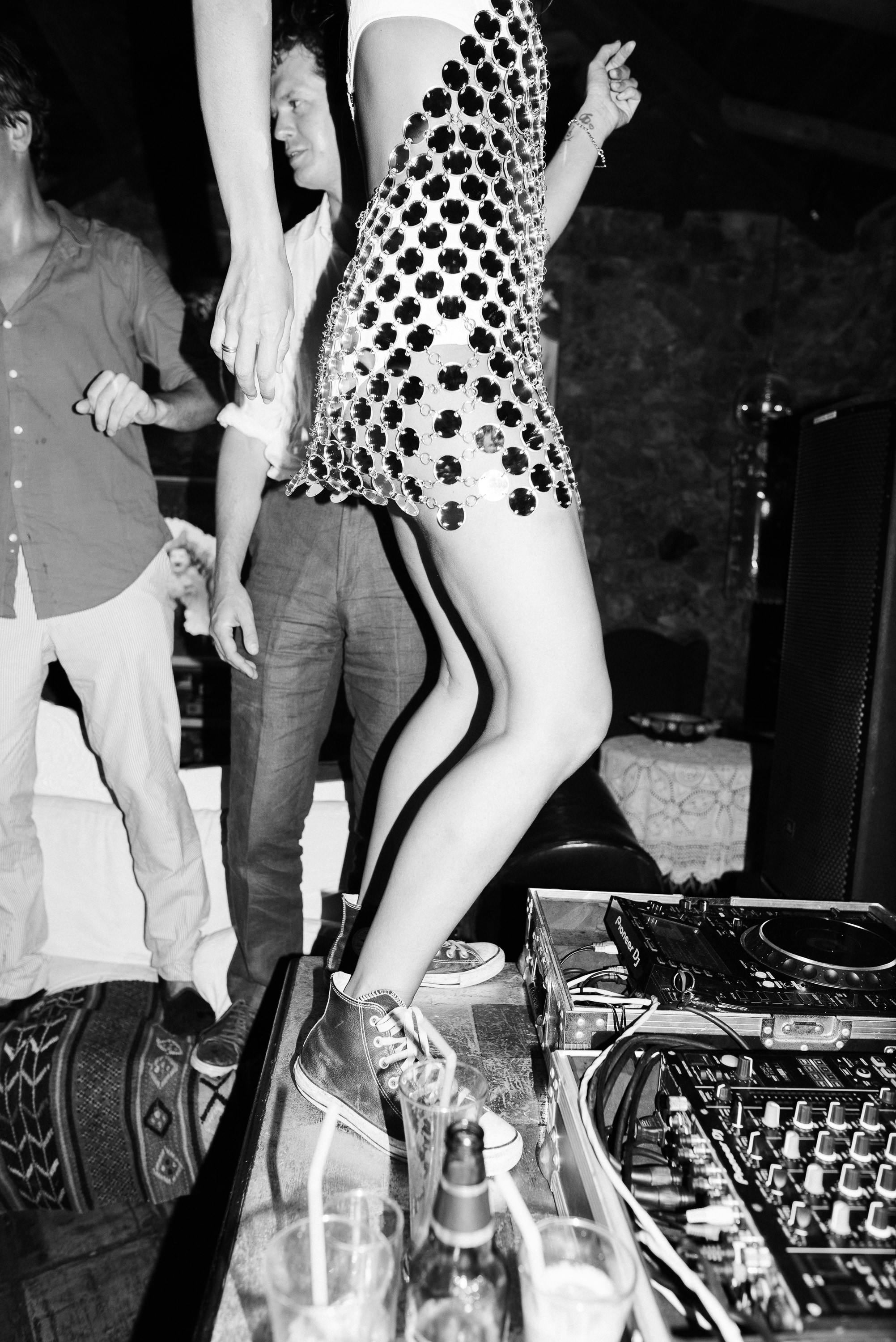 Black and white photo of person dancing energetically on a DJ booth table wearing patterned outfit and white sneakers. DJ equipment is visible below, with bottles scattered on the surface. Another person in white stands nearby in this lively nighttime wedding reception moment.