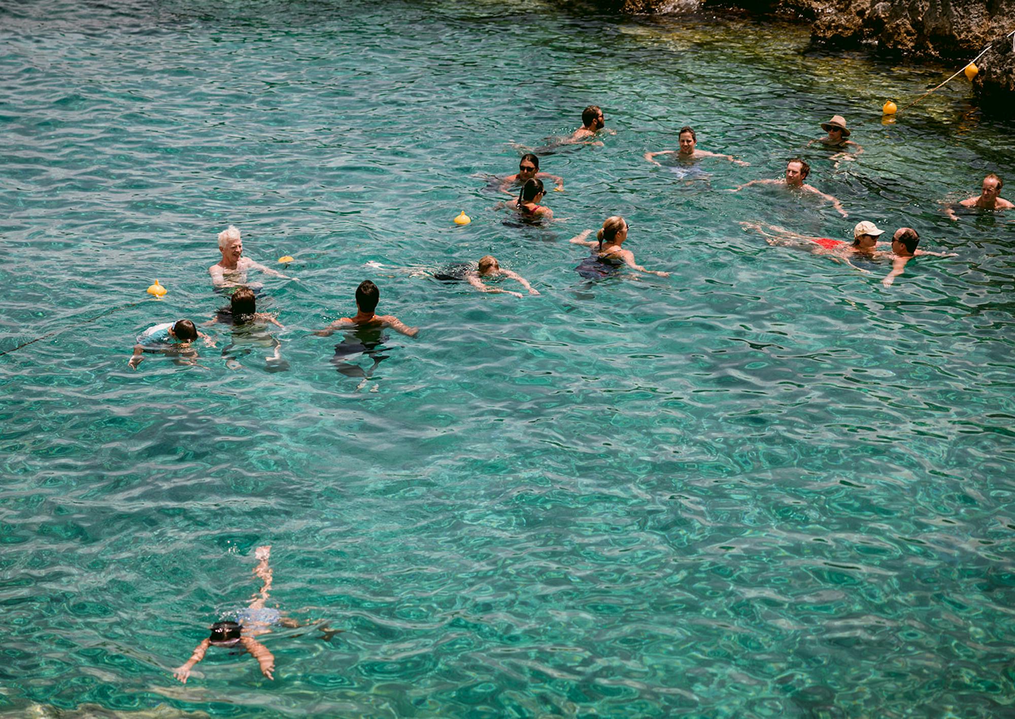 Group of guests swimming together in crystal-clear turquoise Mediterranean waters. Multiple swimmers are scattered across the calm sea near rocky coastline, enjoying a refreshing group swim during the destination celebration.