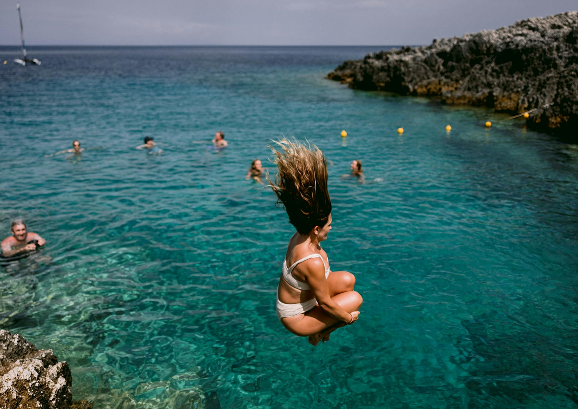 Person jumping into crystal-clear turquoise Mediterranean waters from rocky coastline, captured mid-air with hair flying upward. Multiple swimmers are visible in the background enjoying the calm, transparent sea under overcast skies.