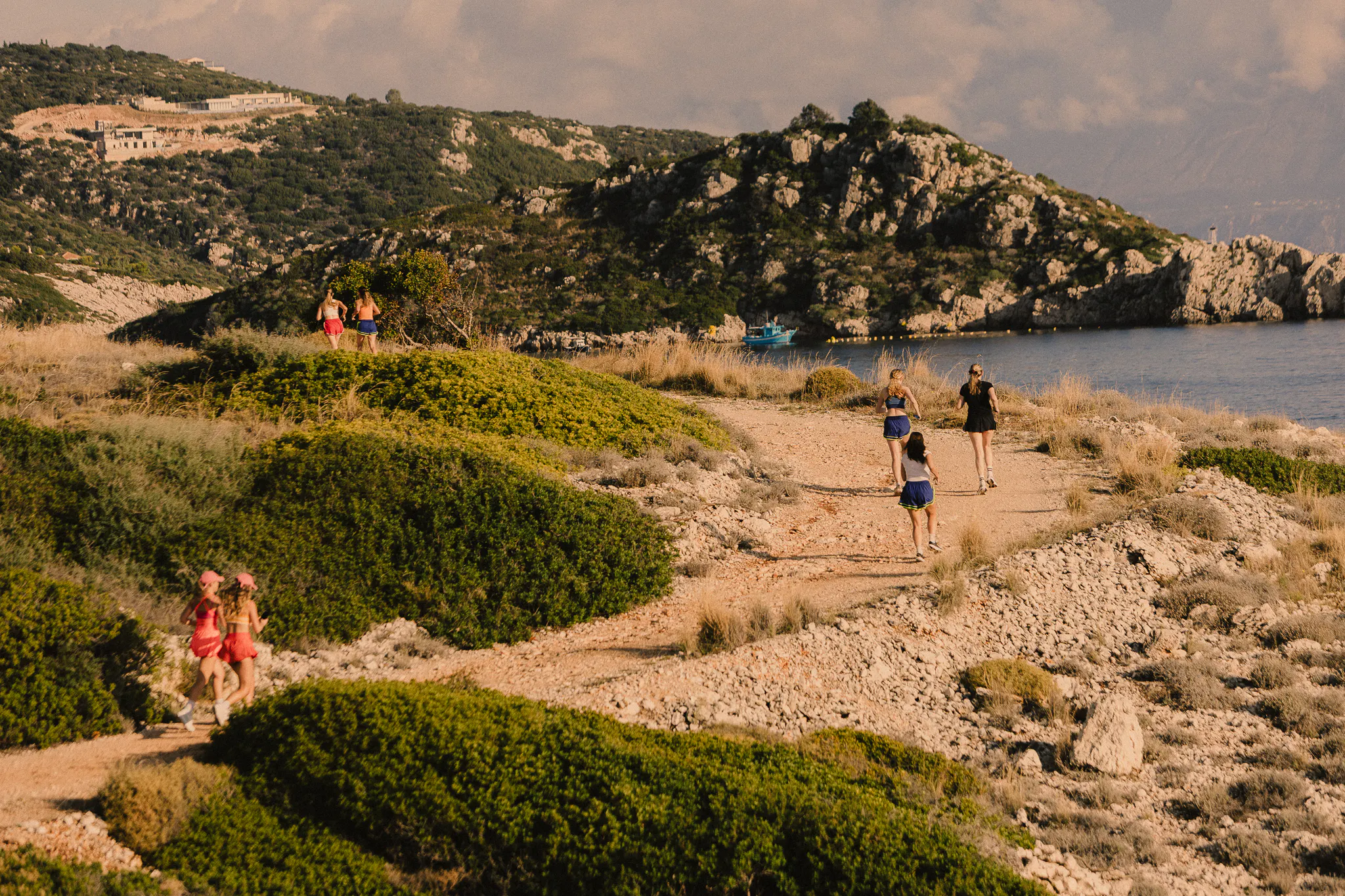 A group of women wearing bright colours and running along a rugged path next to the sea on a bright, sunny day.