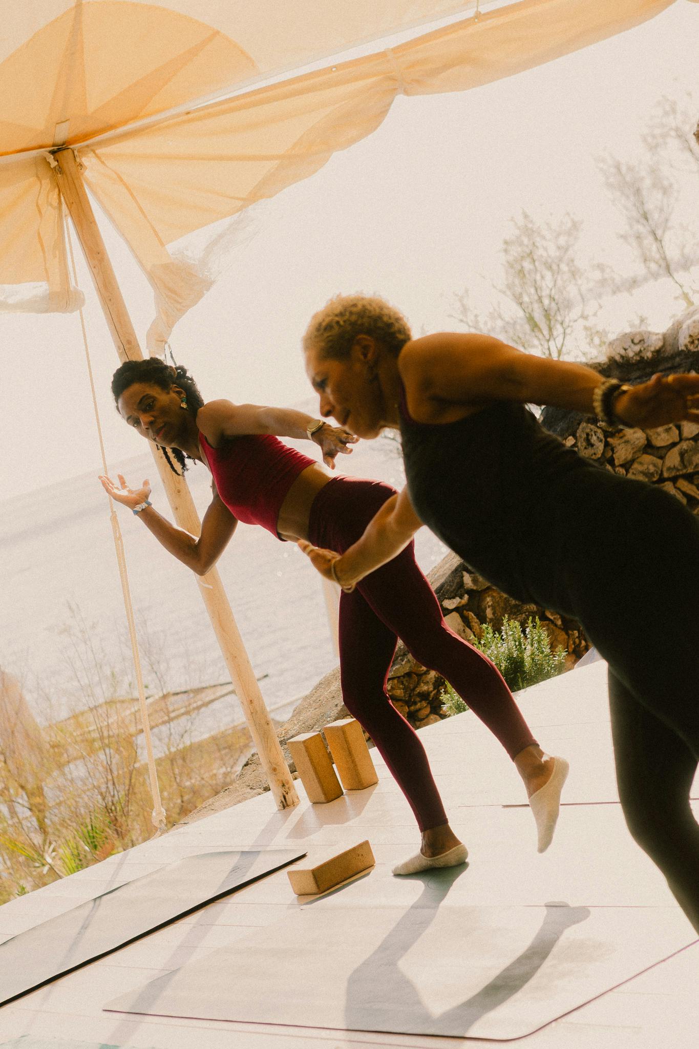 Two women lean in yoga poses under a tented roof with trees and the sea in the background.