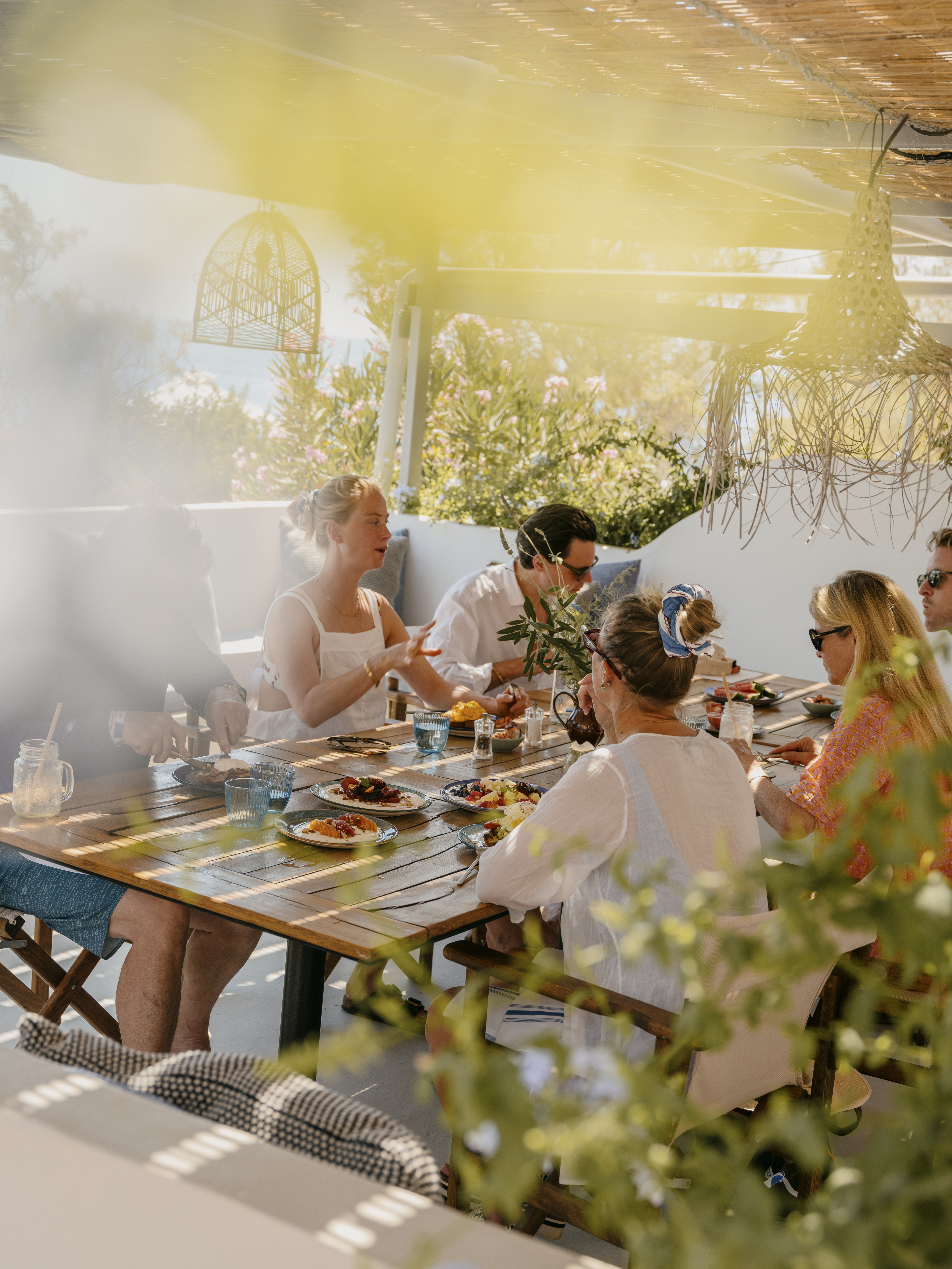 A group of men and women sit around a table of food and drink under dappled sunlight.