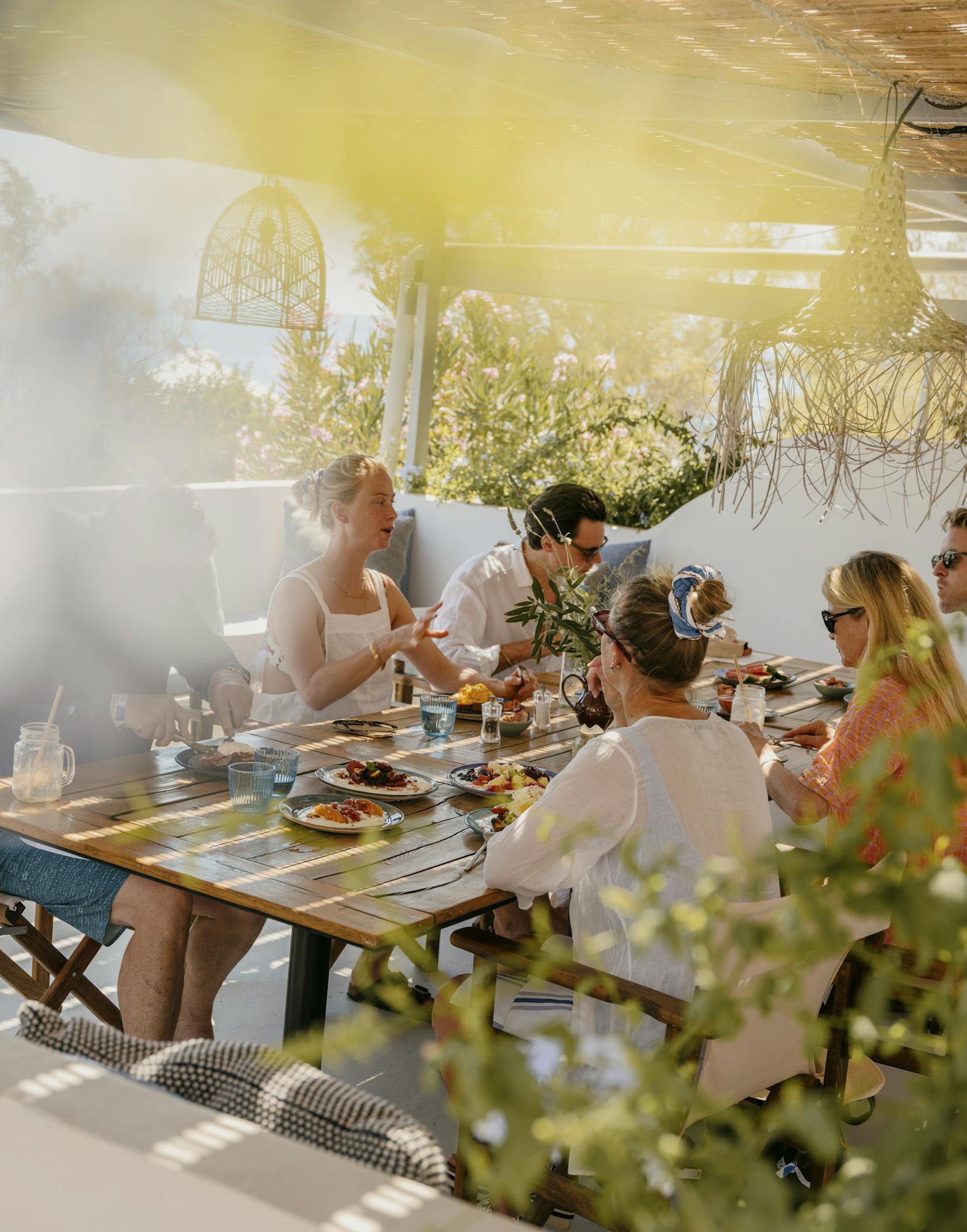 A group of men and women sit around a table of food and drink under dappled sunlight.