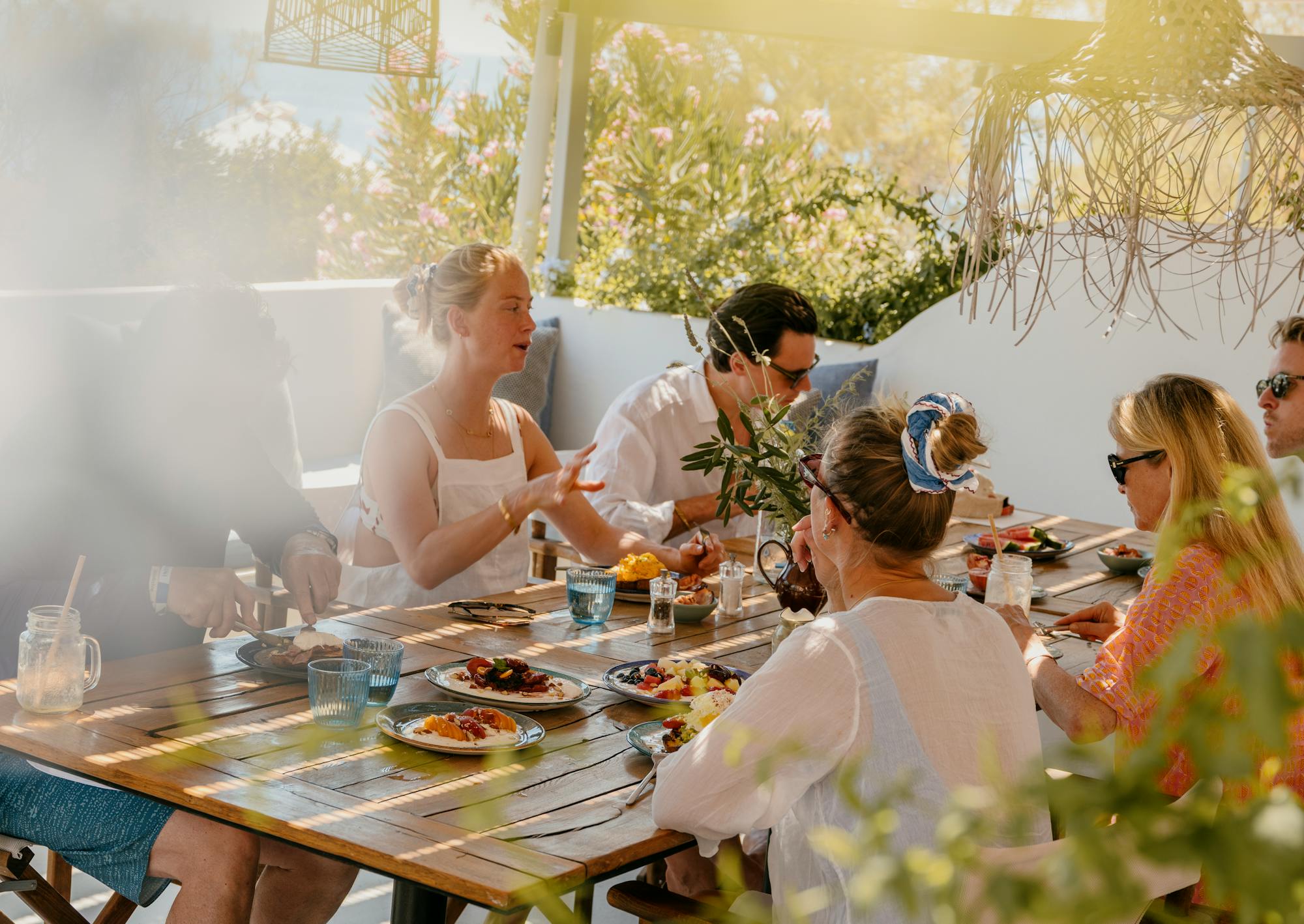 A group of men and women sit around a table of food and drink under dappled sunlight.