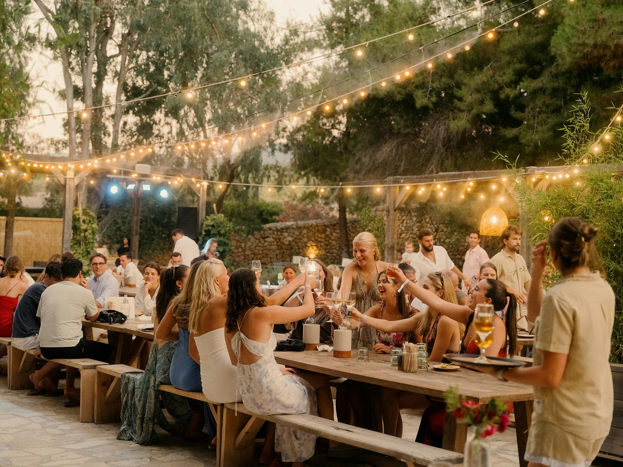 Guests raising glasses in a celebratory toast at an outdoor dinner party under string lights at golden hour, creating a warm, joyful atmosphere.
