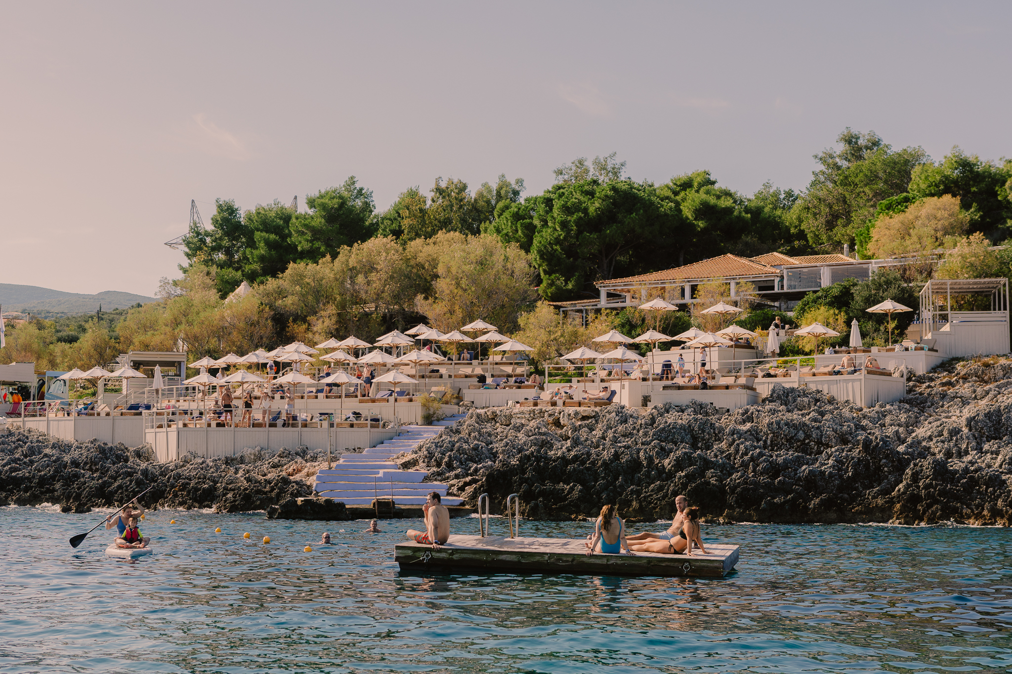 Coastal Mediterranean resort with swimmers and sunbathers enjoying turquoise waters, terraced seating areas with umbrellas, and hillside buildings amid greenery under soft evening light.