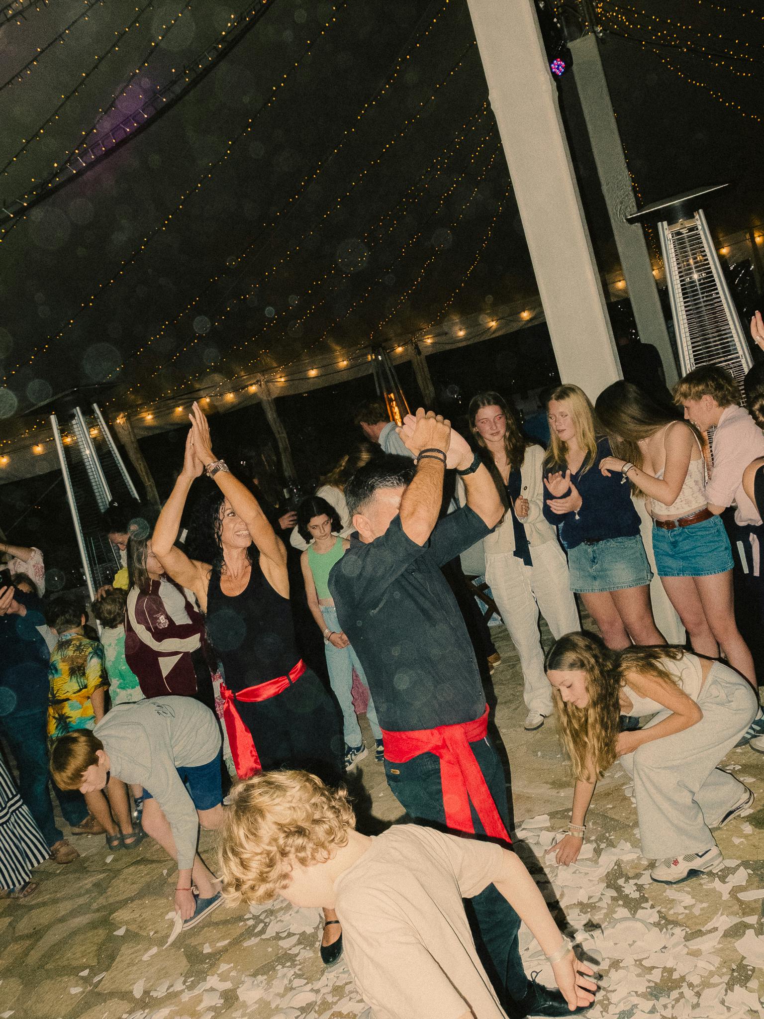 Guests dance energetically under string lights at night, some wearing traditional Greek costume with red sashes, while broken plates litter the floor—the joyful chaos of a Greek celebration in full swing.