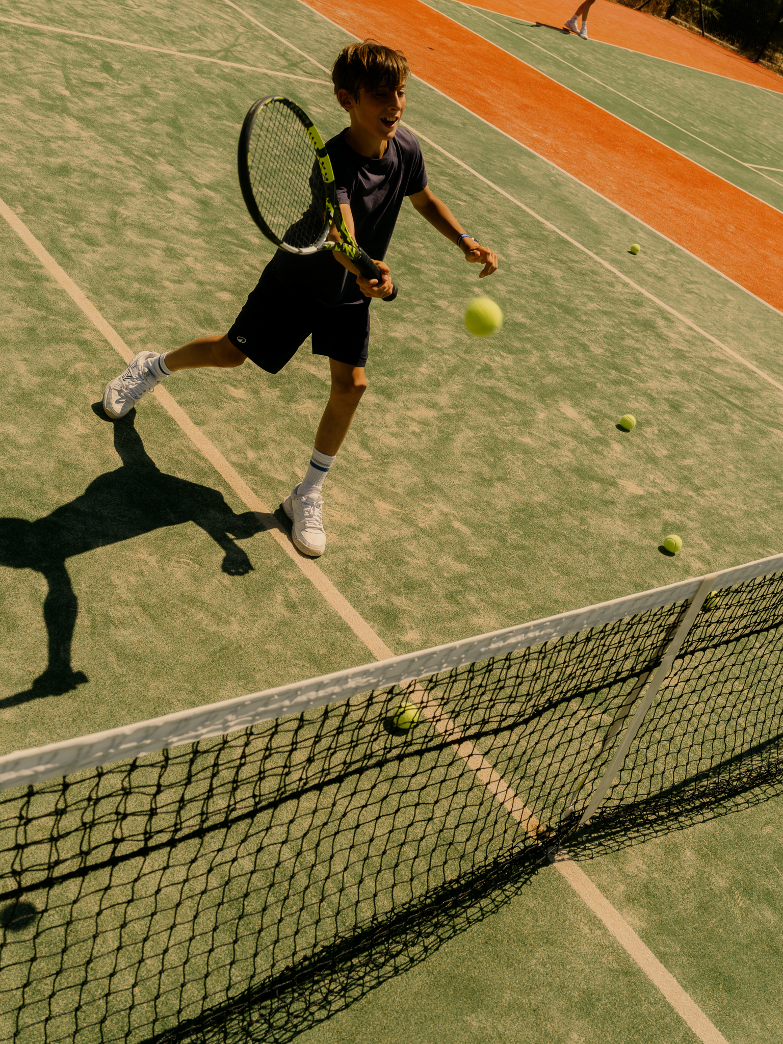A young player mid-swing returns a shot on a sun-drenched tennis court, demonstrating focused athletic engagement in the resort's sports programming.