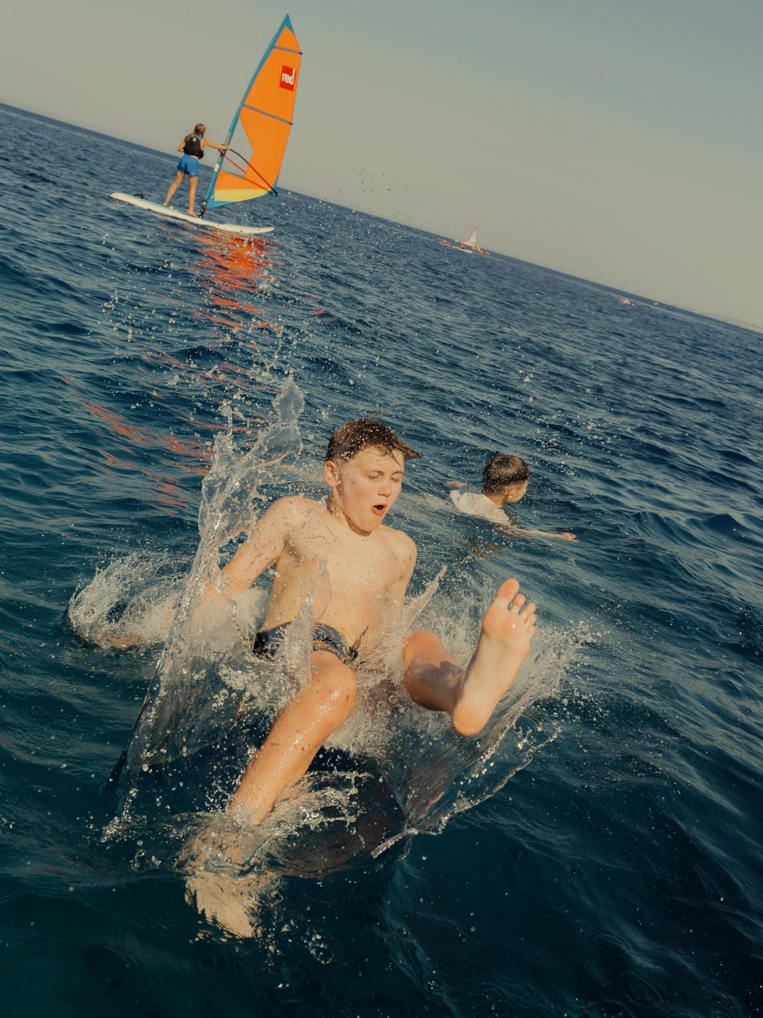 A child creates a dramatic splash while jumping into dark blue sea water, with another windsurfing on an orange-sailed board in the background—capturing the exhilarating variety of water sports activities.