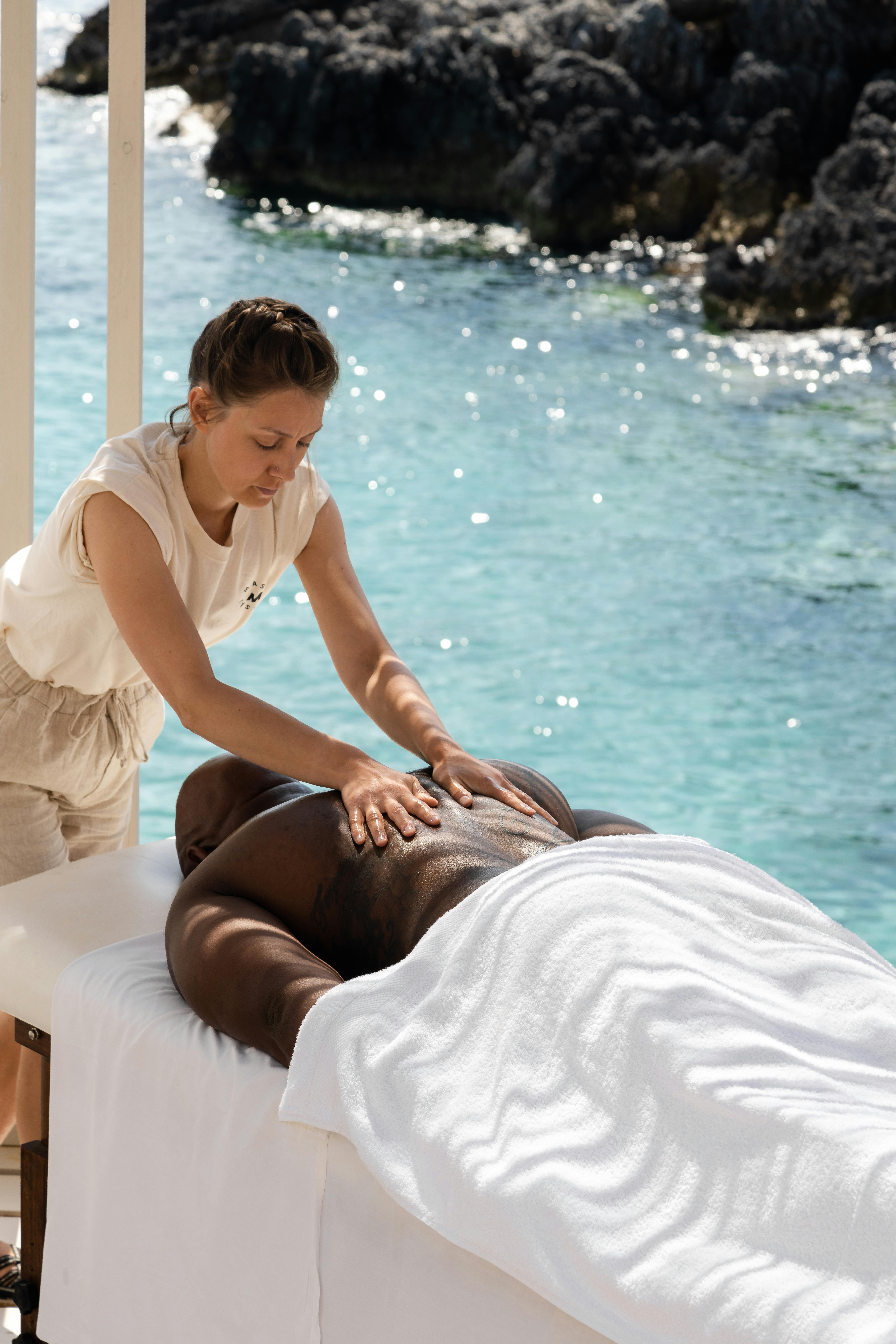 A massage therapist performs a shoulder and upper back massage on a client lying face-down on an outdoor massage table. The therapist wears a white sleeveless top, and the client is draped in white linens. The setting is beside a sparkling turquoise ocean with dark rocky outcrops visible in the background.