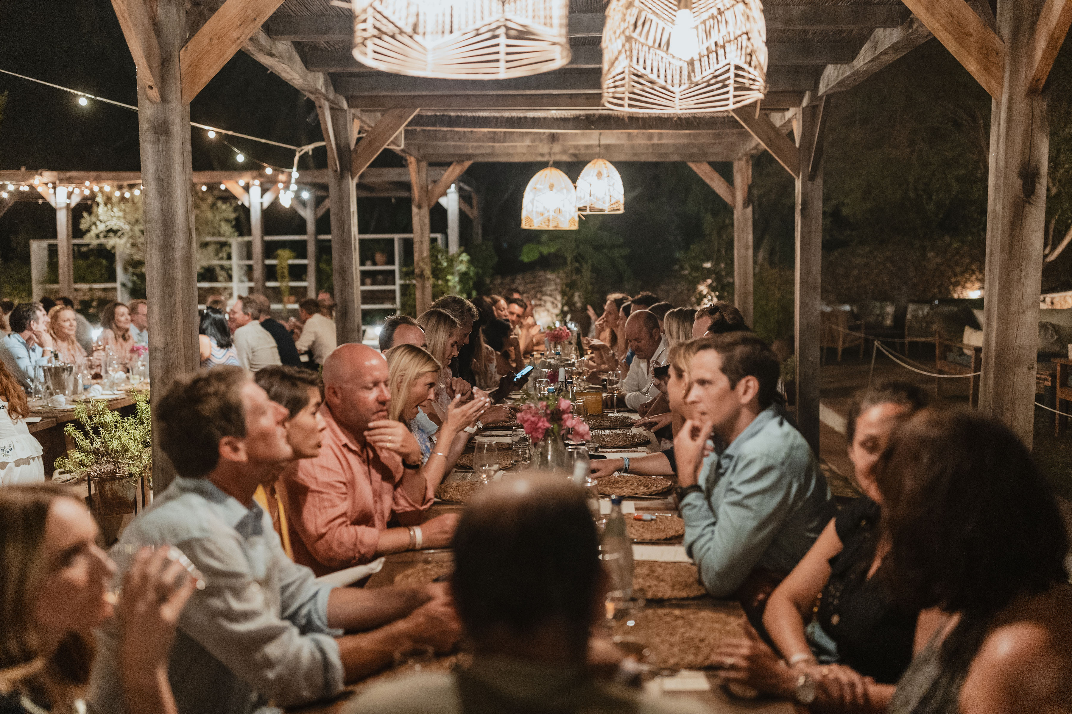 Guests dine together at long communal tables under a wooden pavilion structure illuminated by string lights and geometric pendant lamps, creating a warm, convivial evening atmosphere for an outdoor celebration.