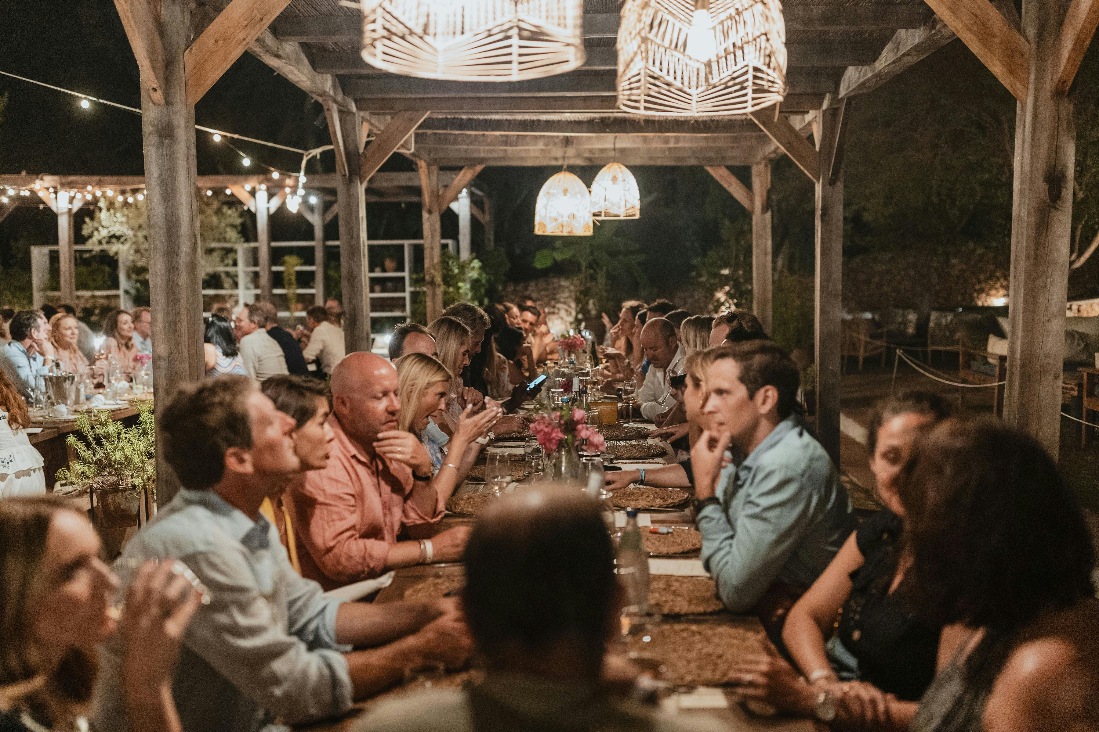 Guests dine together at long communal tables under a wooden pavilion structure illuminated by string lights and geometric pendant lamps, creating a warm, convivial evening atmosphere for an outdoor celebration.
