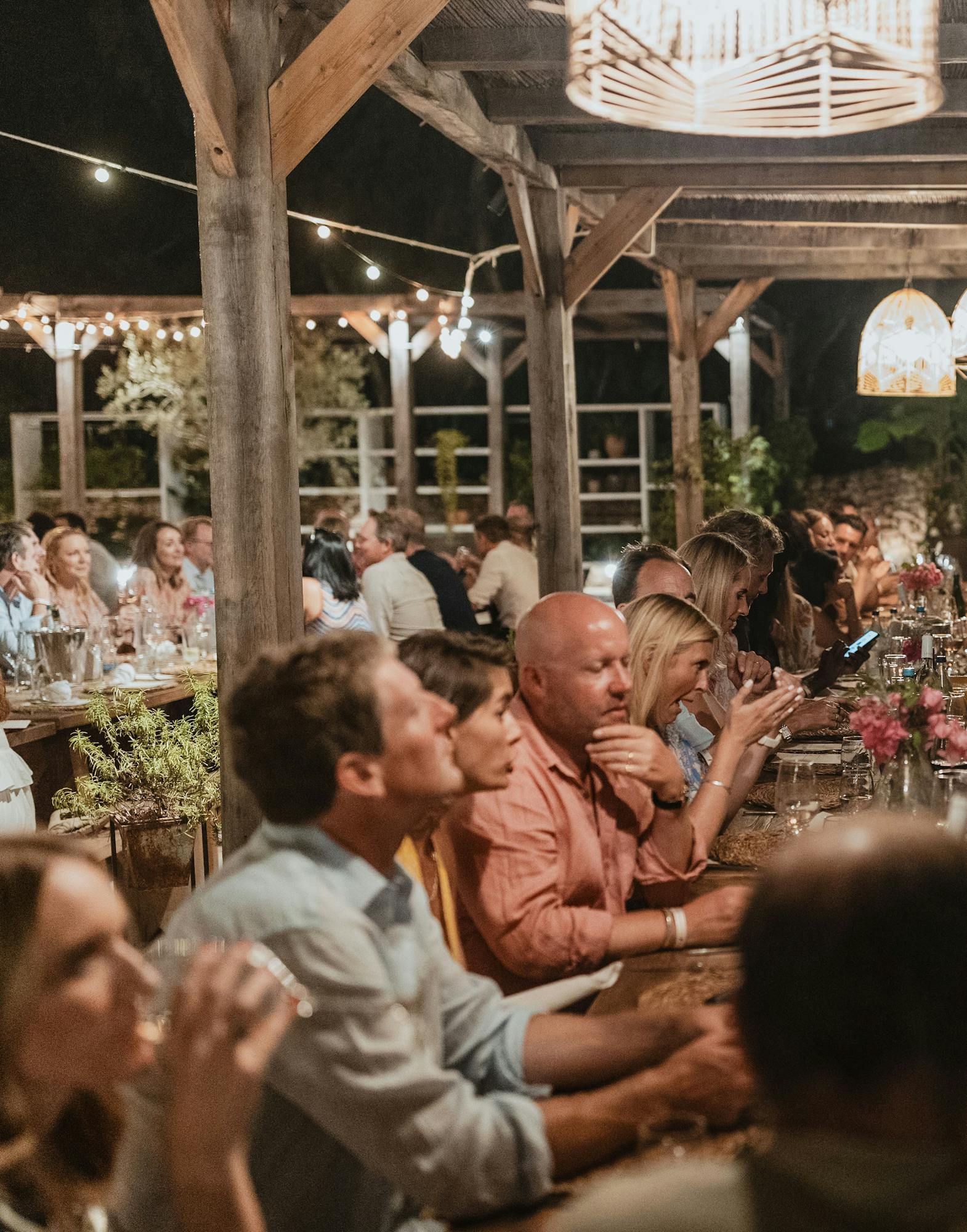 Guests dine together at long communal tables under a wooden pavilion structure illuminated by string lights and geometric pendant lamps, creating a warm, convivial evening atmosphere for an outdoor celebration.