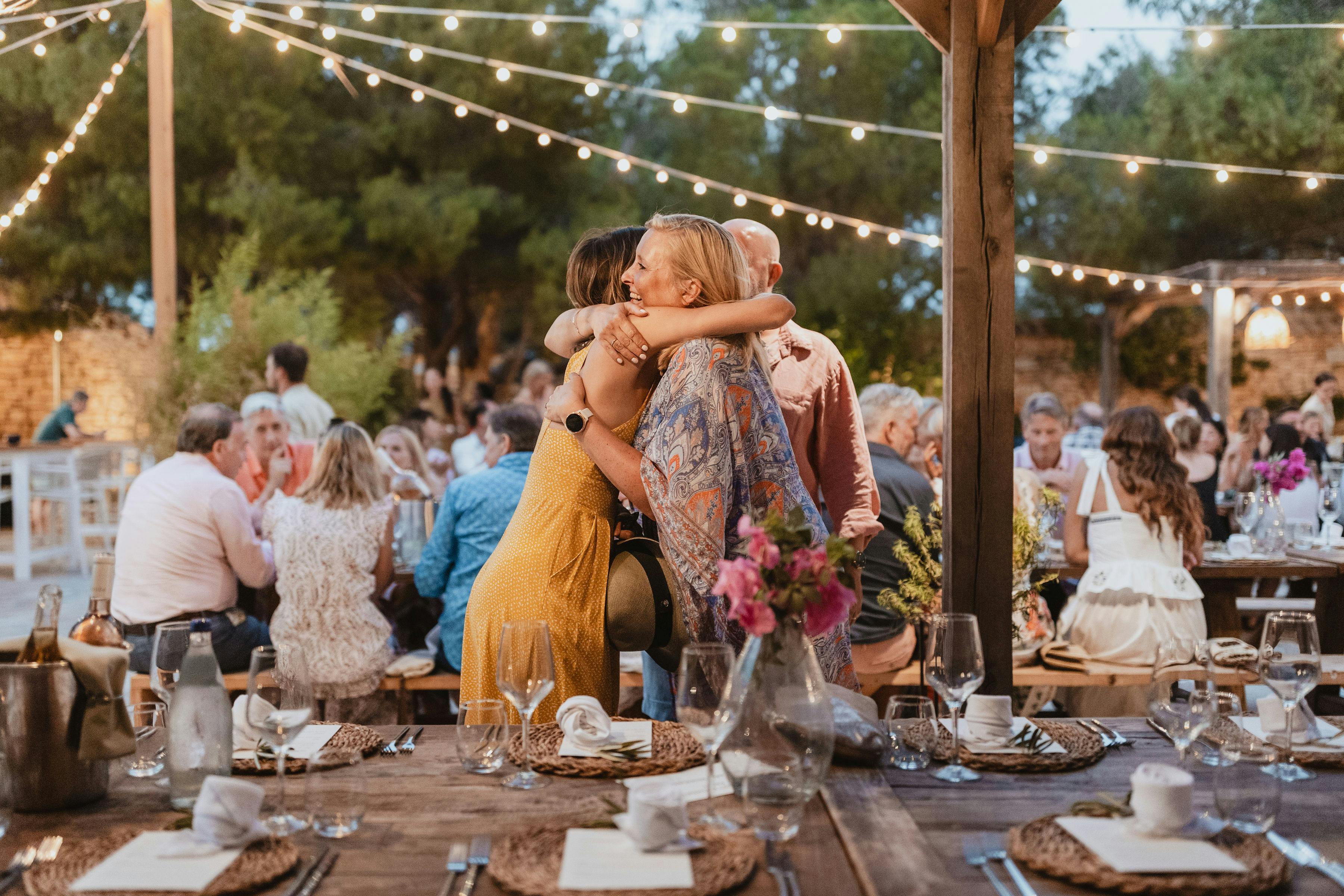 Two women embrace warmly at an outdoor dinner celebration under string lights, surrounded by guests at long wooden tables with rustic place settings and fresh flowers, creating an intimate and joyful atmosphere amid trees and natural sunlight.