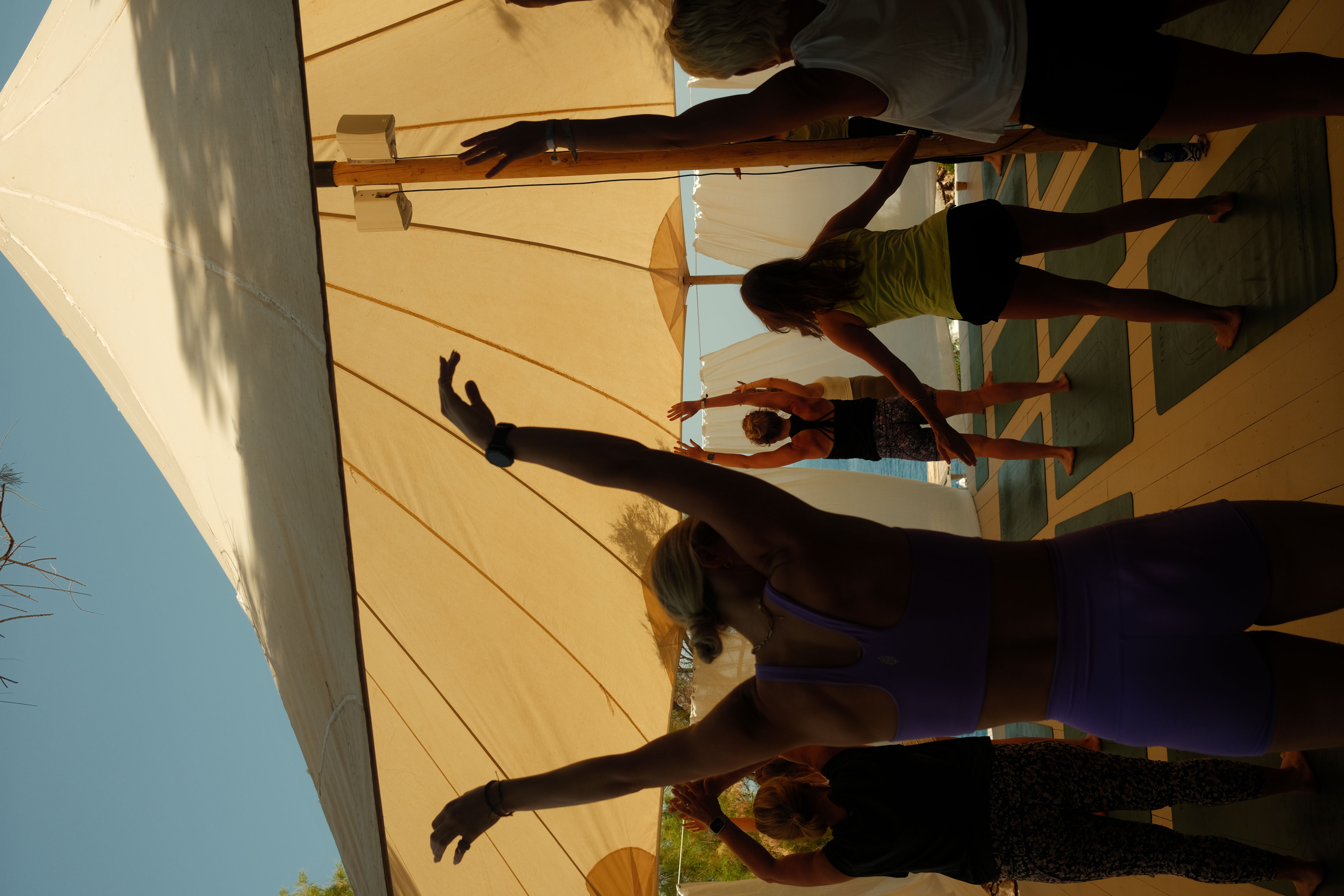 Silhouettes of people doing yoga with arms raised overhead on mats under a beige canopy, backlit by golden sunlight with the sea visible in the background.