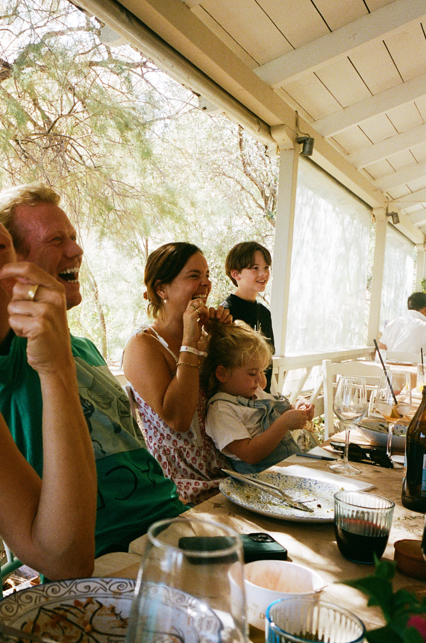 Family dining together at an outdoor restaurant table under a white pergola with natural light and greenery.