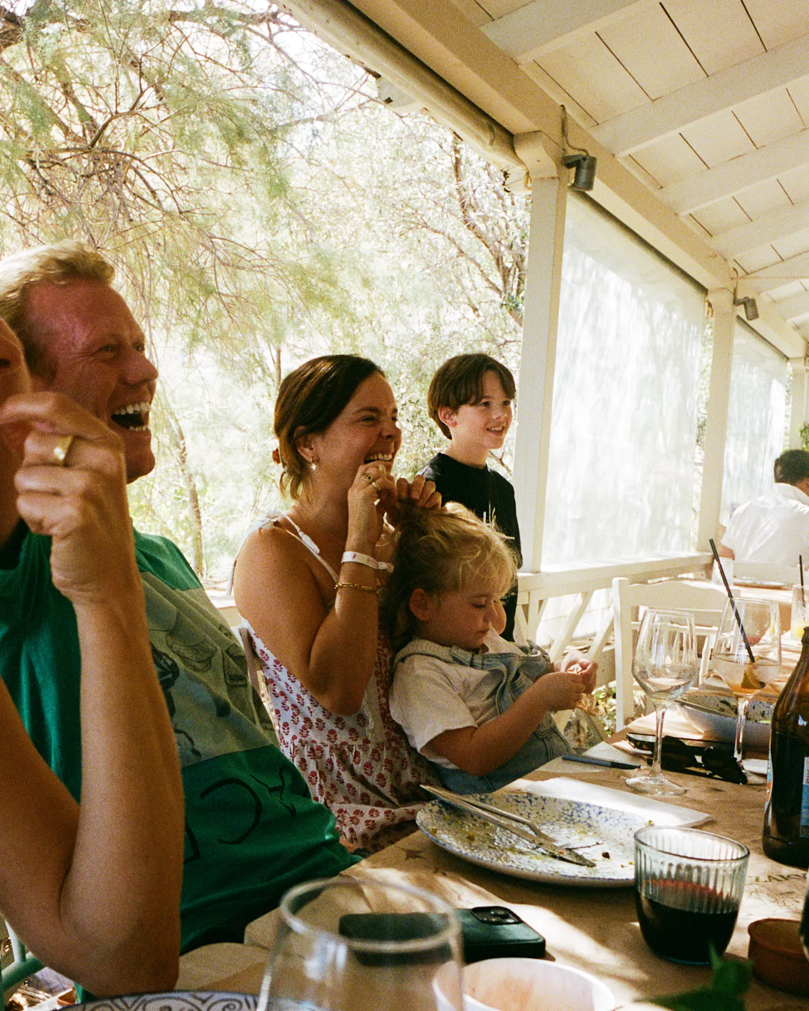 Family dining together at an outdoor restaurant table under a white pergola with natural light and greenery.