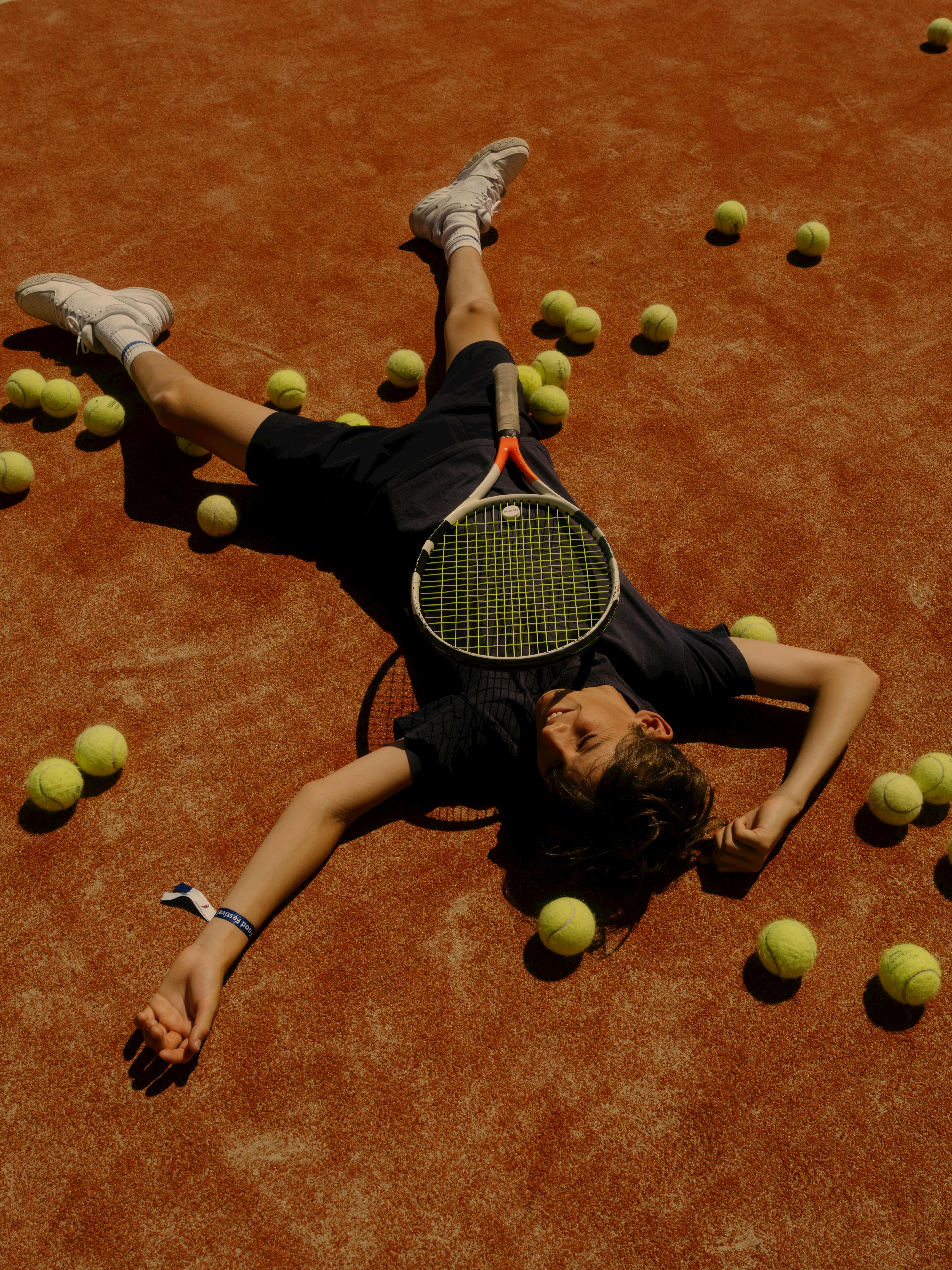 A person lying on a red clay tennis court surrounded by tennis balls, holding a tennis racket, shot from above.