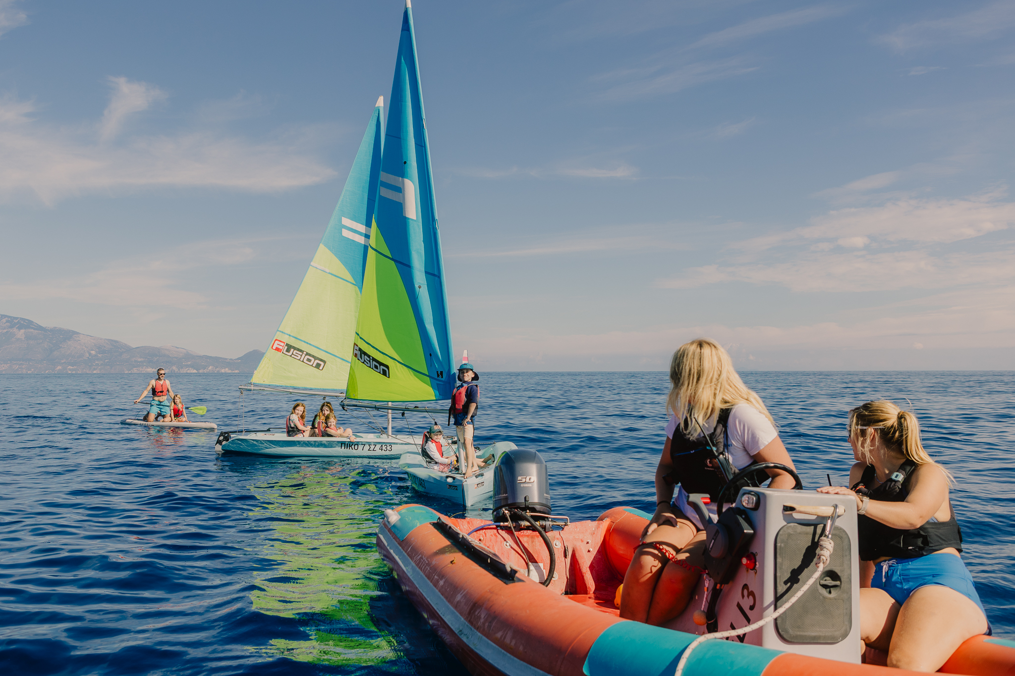 A group of children enjoying a sailing lesson on a calm blue sea, with sailboats and inflatable boats under a clear sky and mountains in the distance.