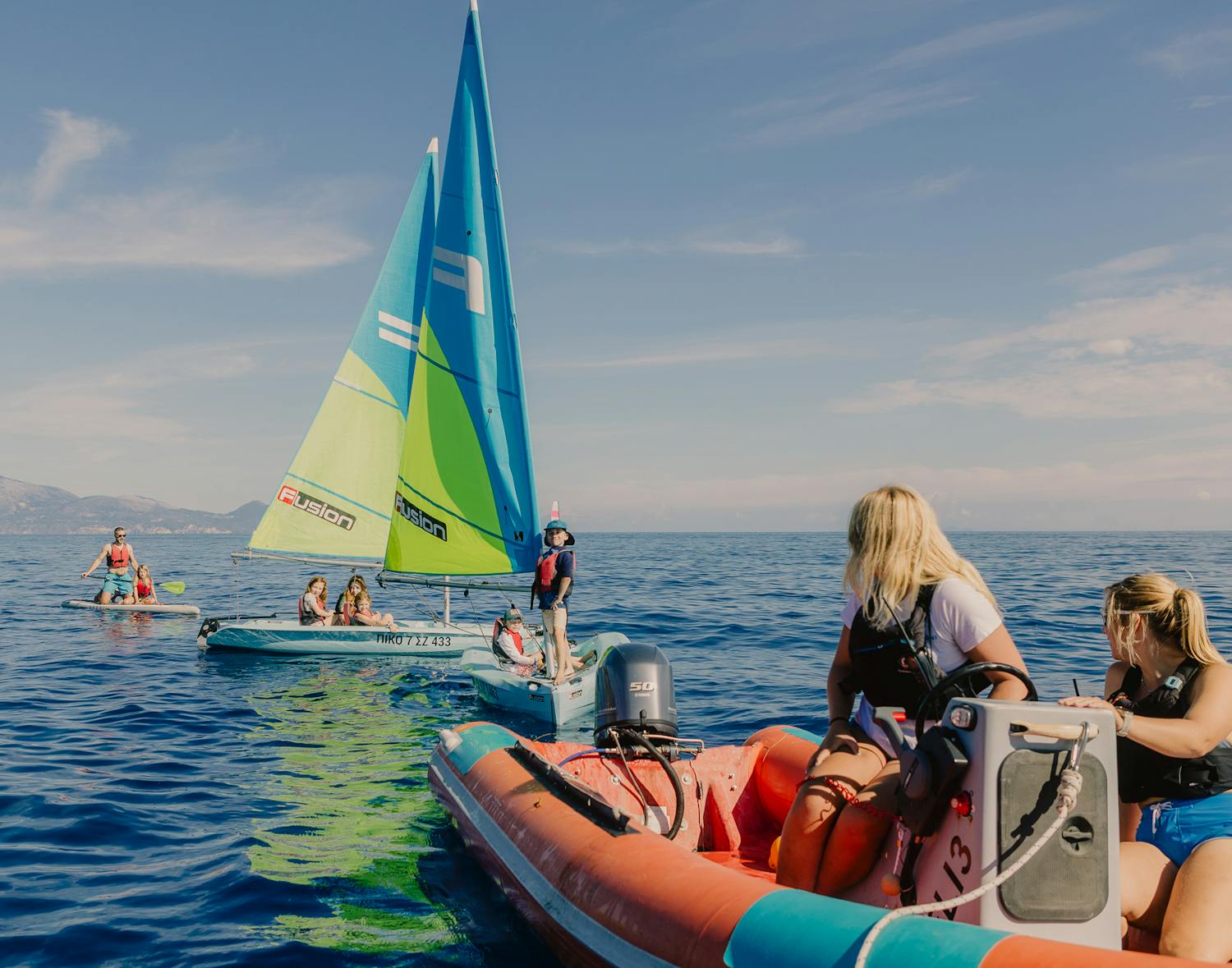 A group of children enjoying a sailing lesson on a calm blue sea, with sailboats and inflatable boats under a clear sky and mountains in the distance.