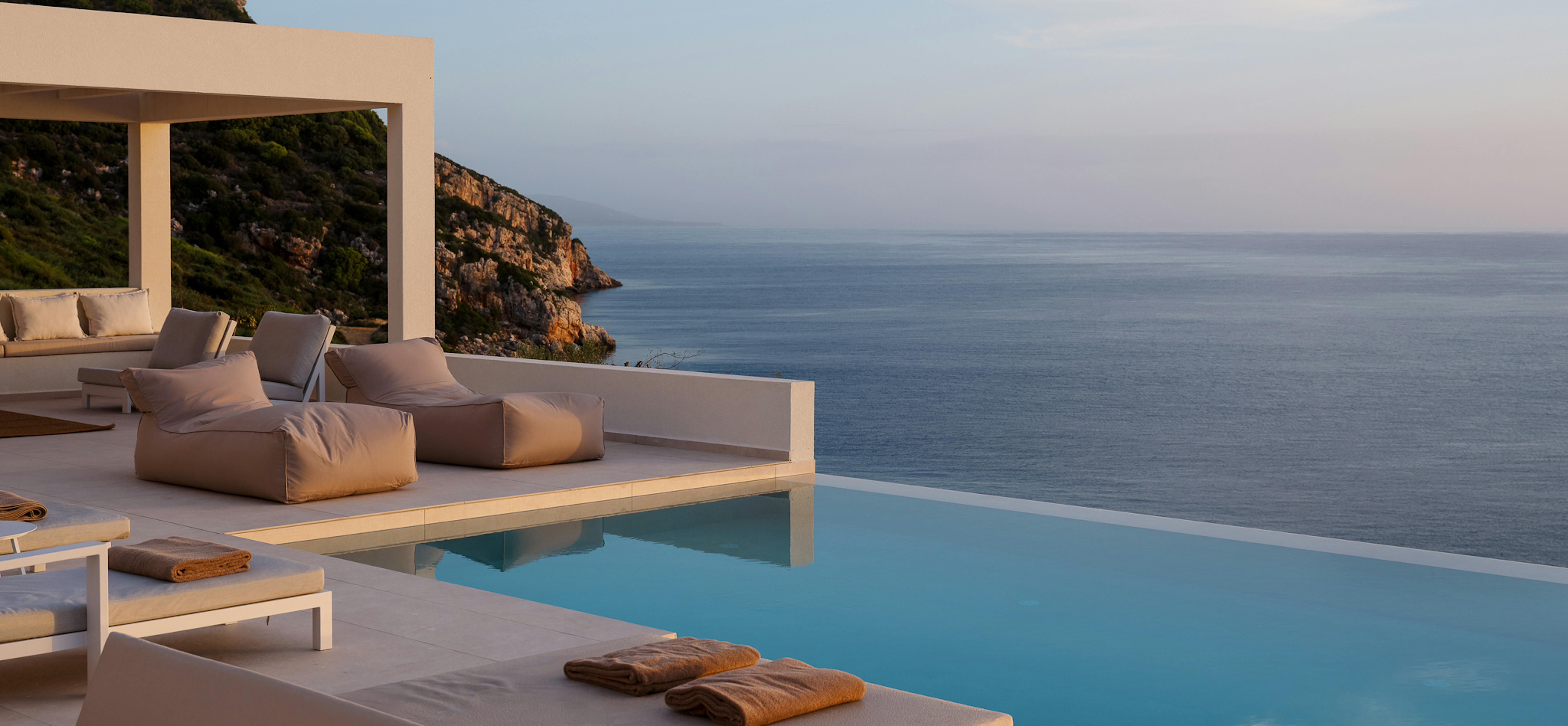 Pool deck at dusk with loungers and cliffside Mediterranean setting.