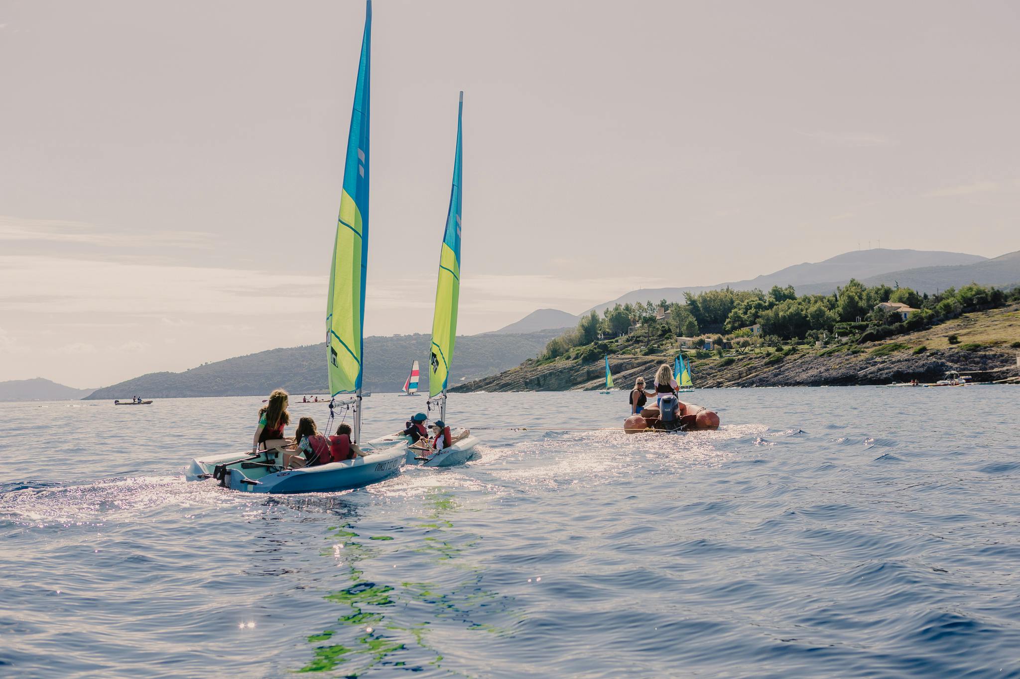 Two sailboats with blue and green sails move through the sea. Each one contains a group of young children in life jackets. They are towed by a speedboat, with a rocky and green landscape in the distance.