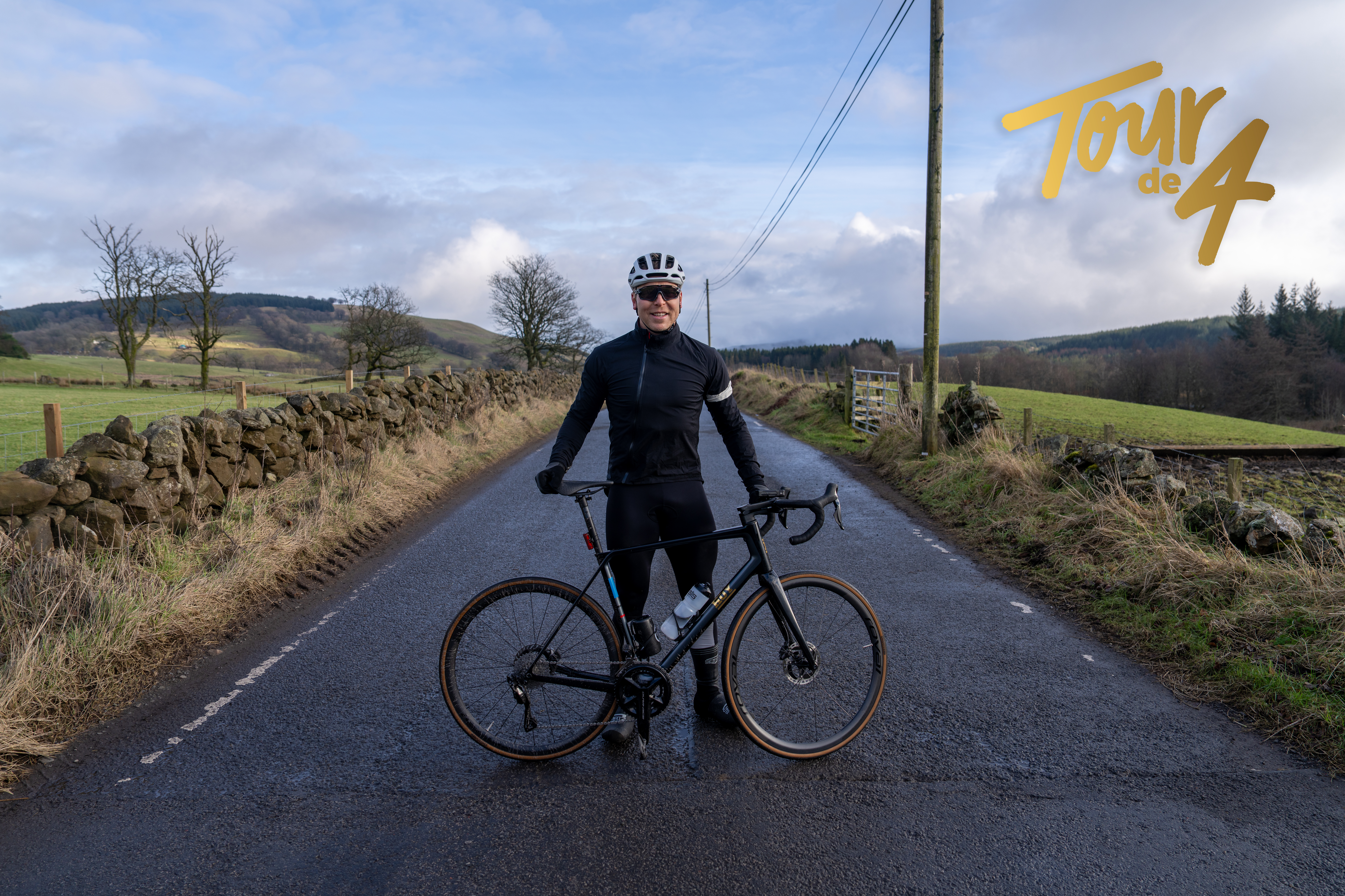 A man in a grey helmet and dark lycra cycle gear stands in the middle of a country lane, holding a bicycle and smiling into the camera.