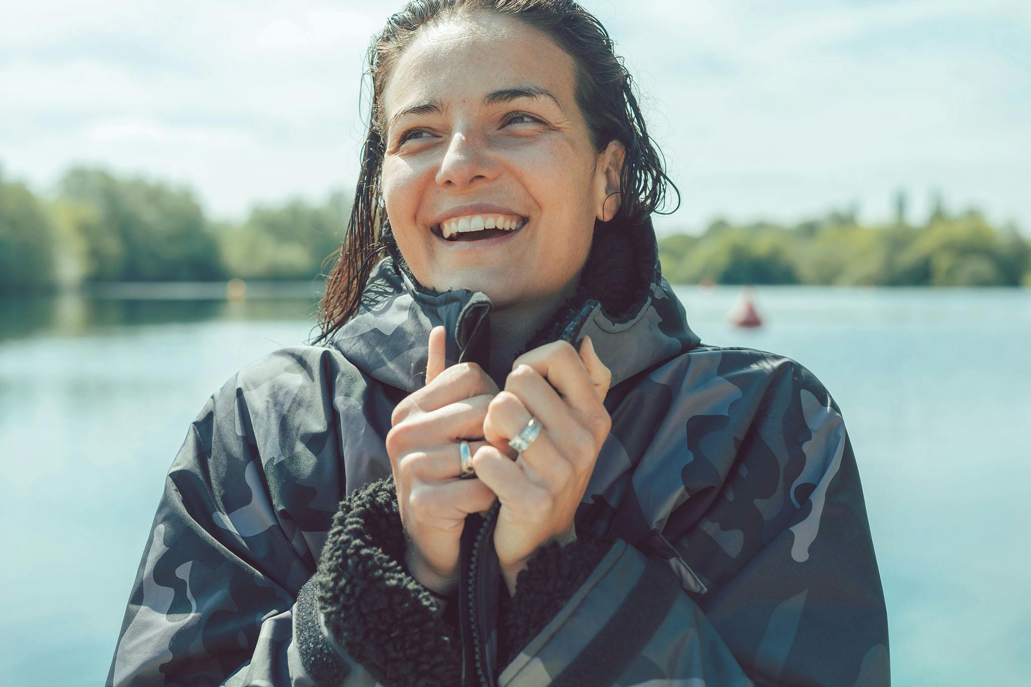 A woman with brown hair wearing a camouflage-print dry robe smiles past the camera, with blurry open water and trees in the background.