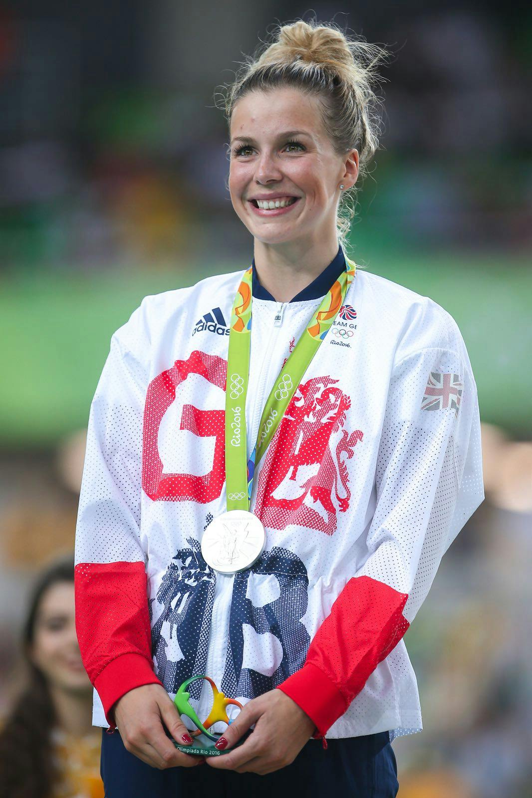 A woman with blonde hair, wearing a white, red and blue top and an olympic silver medal smiles past the camera.