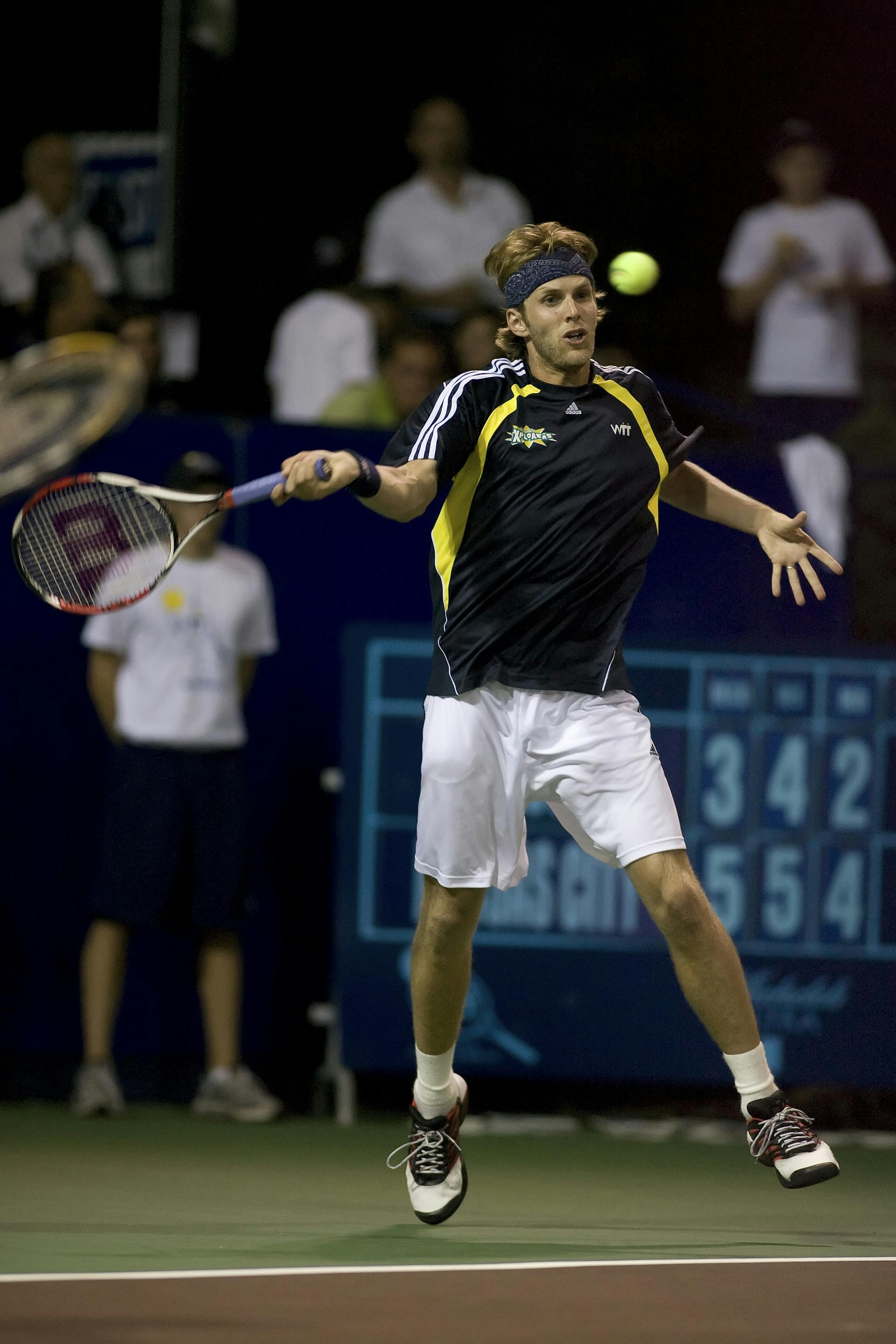 A man in a black top and white shorts, with a sweat band around his head, jumps in the air to hit a tennis ball with his tennis racket. A ball boy and spectators watch in the background.