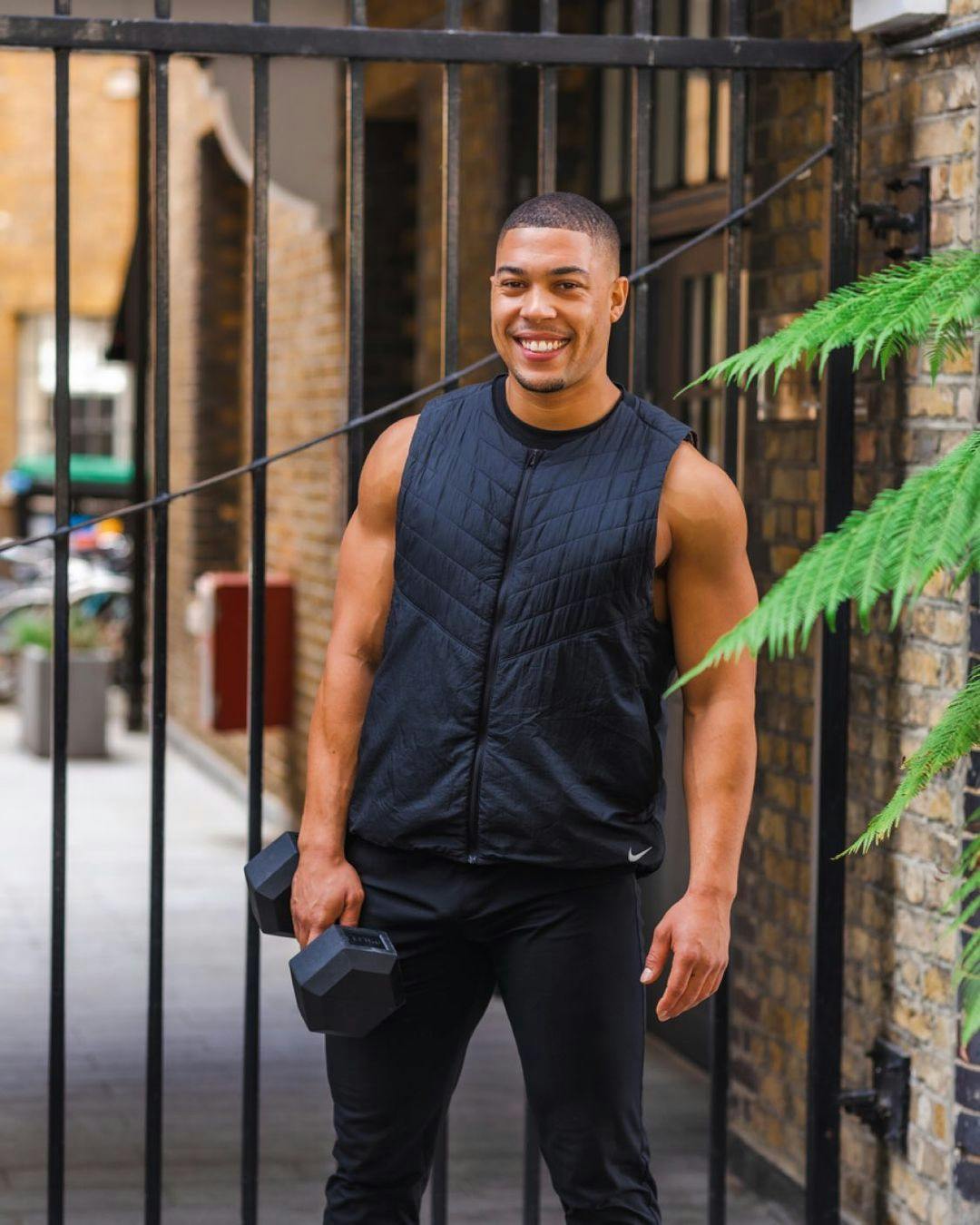 A man in a black vest and black trousers stands in front of a gate holding a weight.
