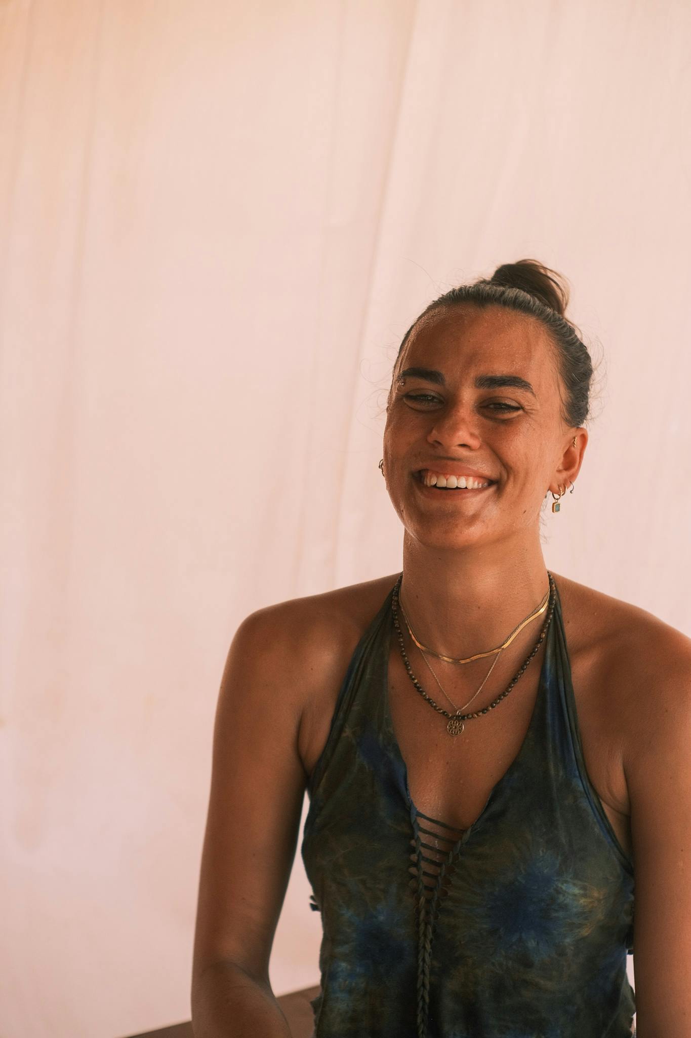 A woman with her hair in a bun laughs with her eyes closed, sat in front of a pale pink curtain.