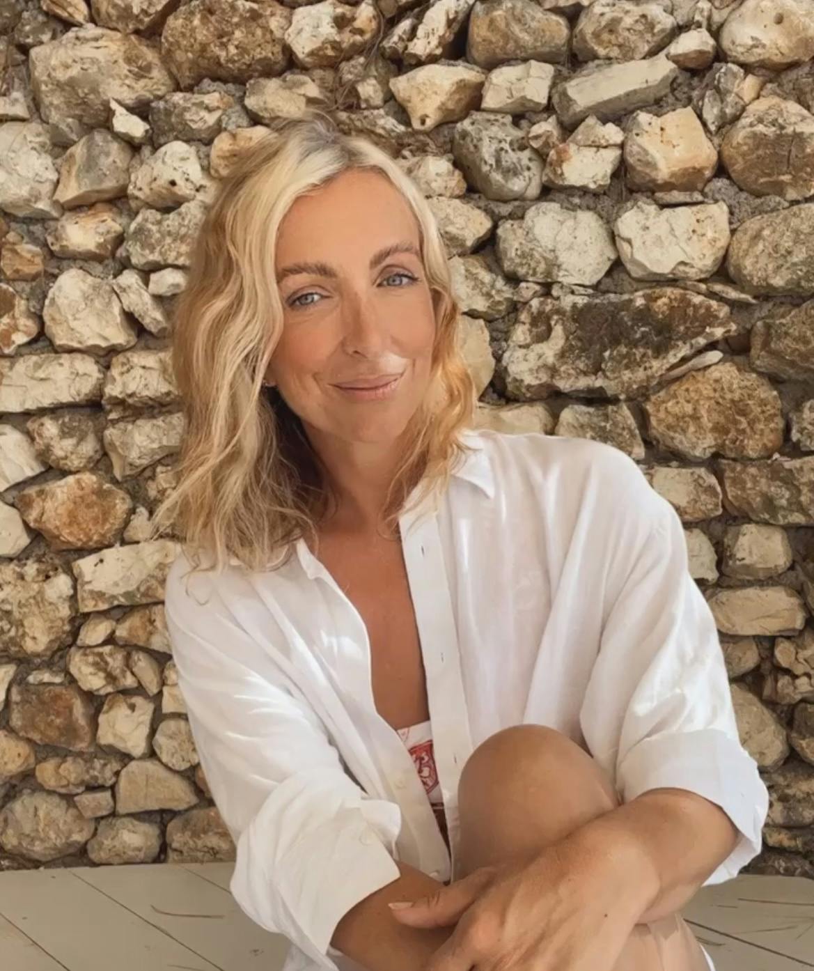 A woman with wavy blonde hair, wearing a white shirt, sits in front of a stone wall, smiling into the camera.