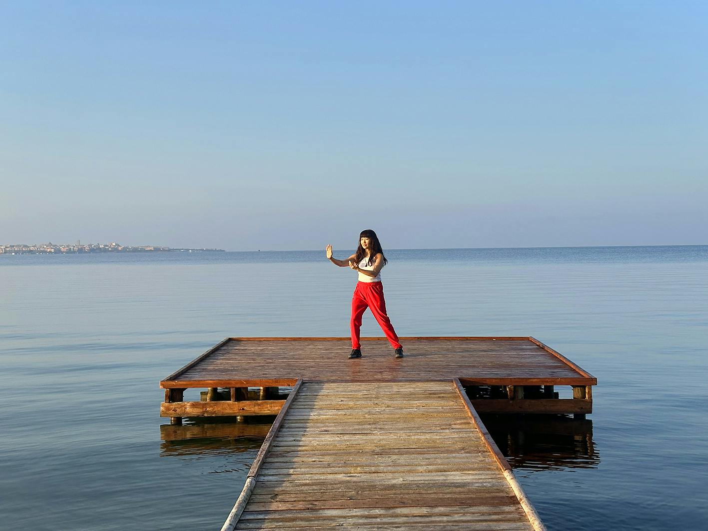 A woman in a white top and red trousers stands on a wooden pontoon, surrounded by blue sea and a clear blue sky, with a cityscape in the far distance.
