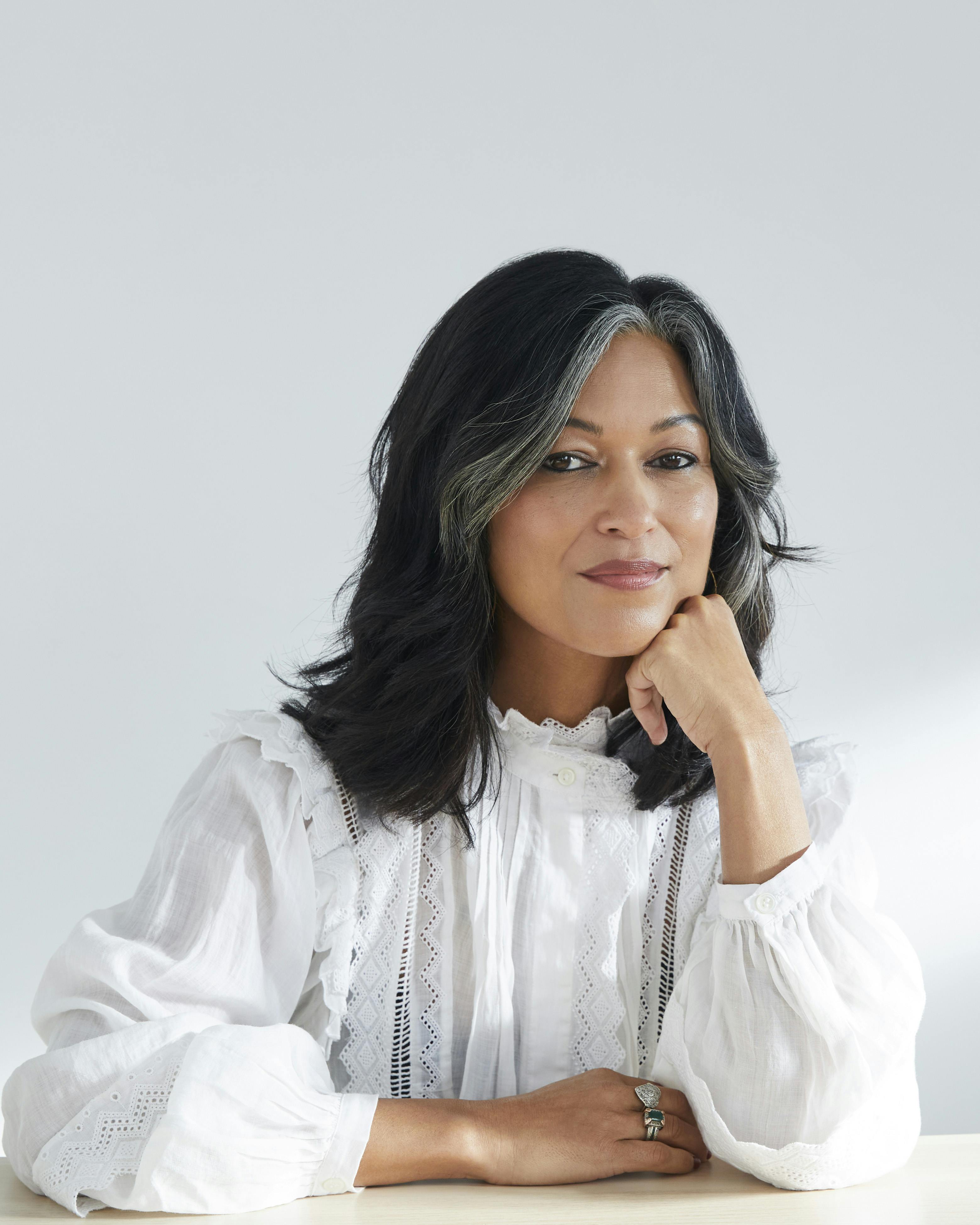 A woman in a white blouse poses for the camera with one hand resting against her chin, the other laid on the desk in front of her.