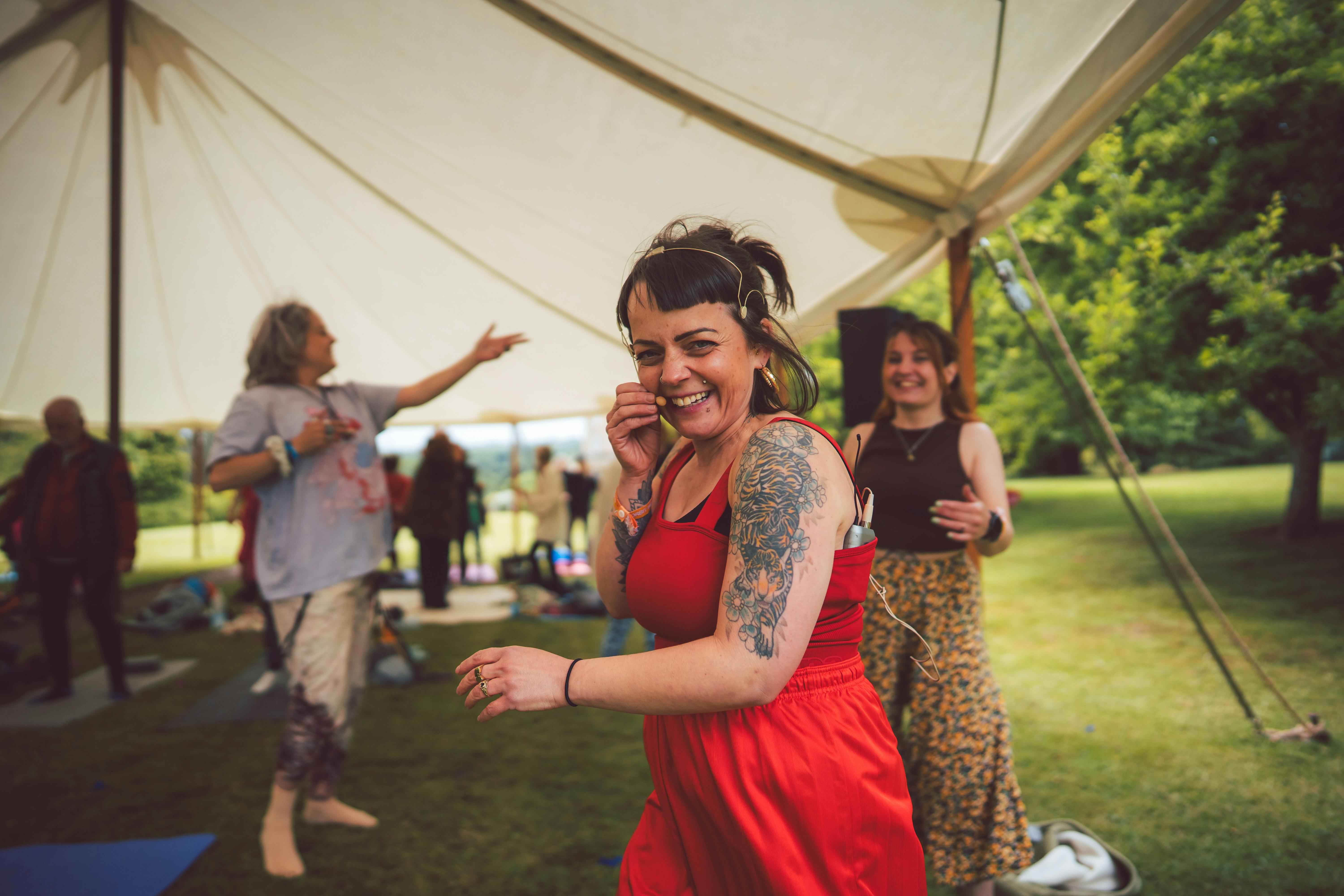 A woman in a red jumpsuit with tattoos on her arm smiles and laughs into the camera. In there background there are other women dancing and laughing, under the cover of a tent in a large green space.