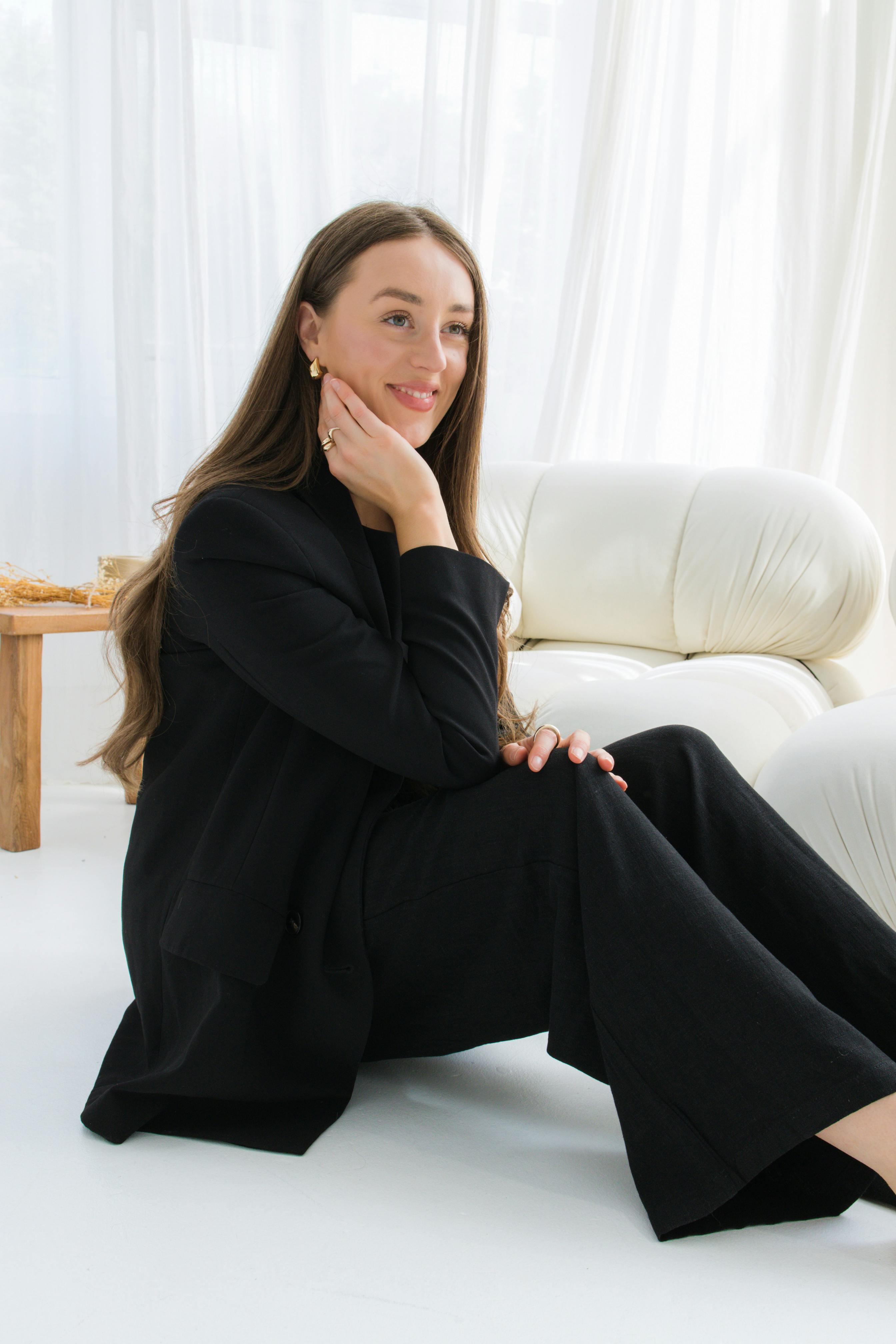 A woman in a black suit sits on the floor with her chin in her hand, with. shite chair and wooden table in the background.