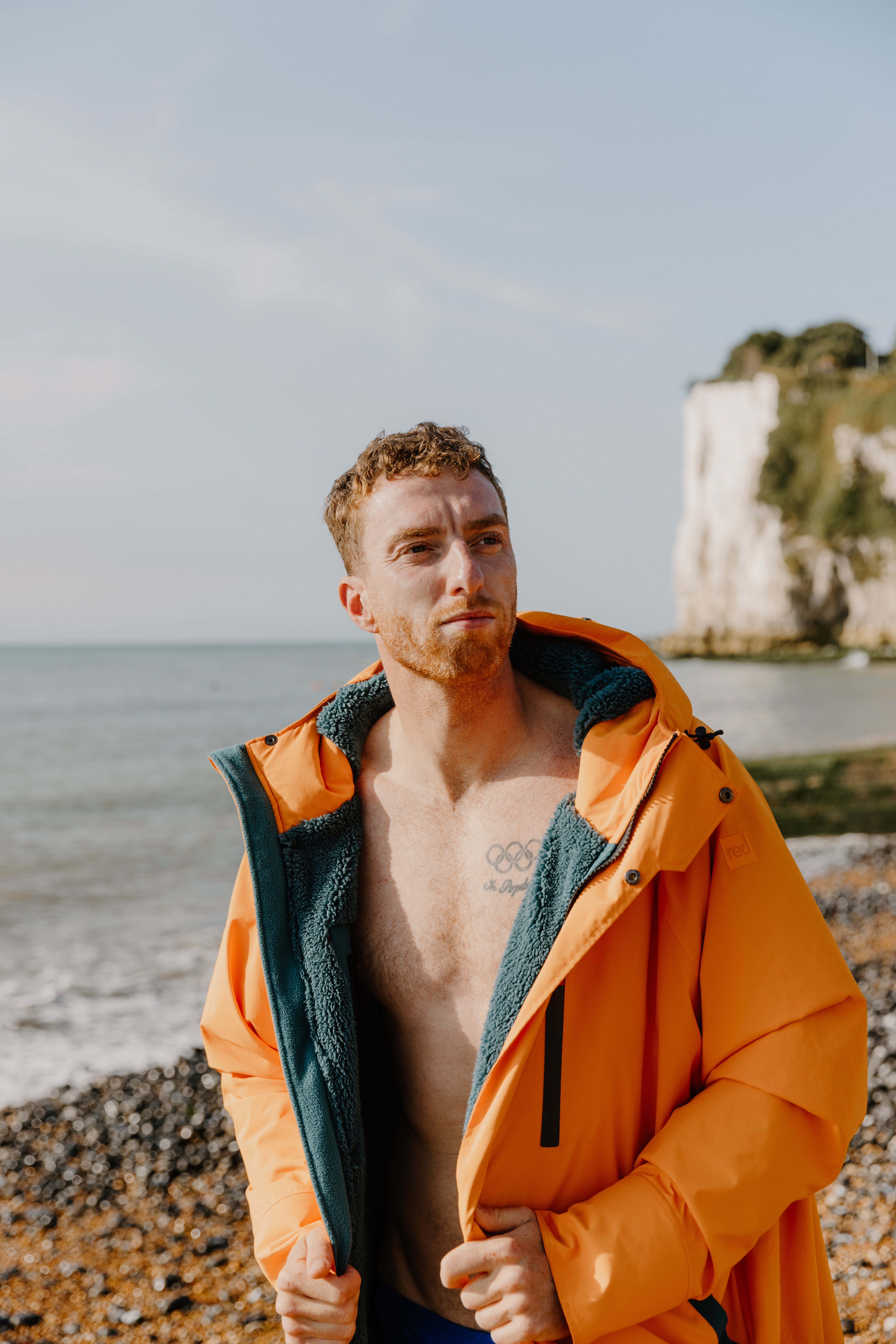 A man standing on a pebble beach wearing an orange parka jacket open to show his bare chest on a backdrop of white cliffs, flat sea and blue skies.