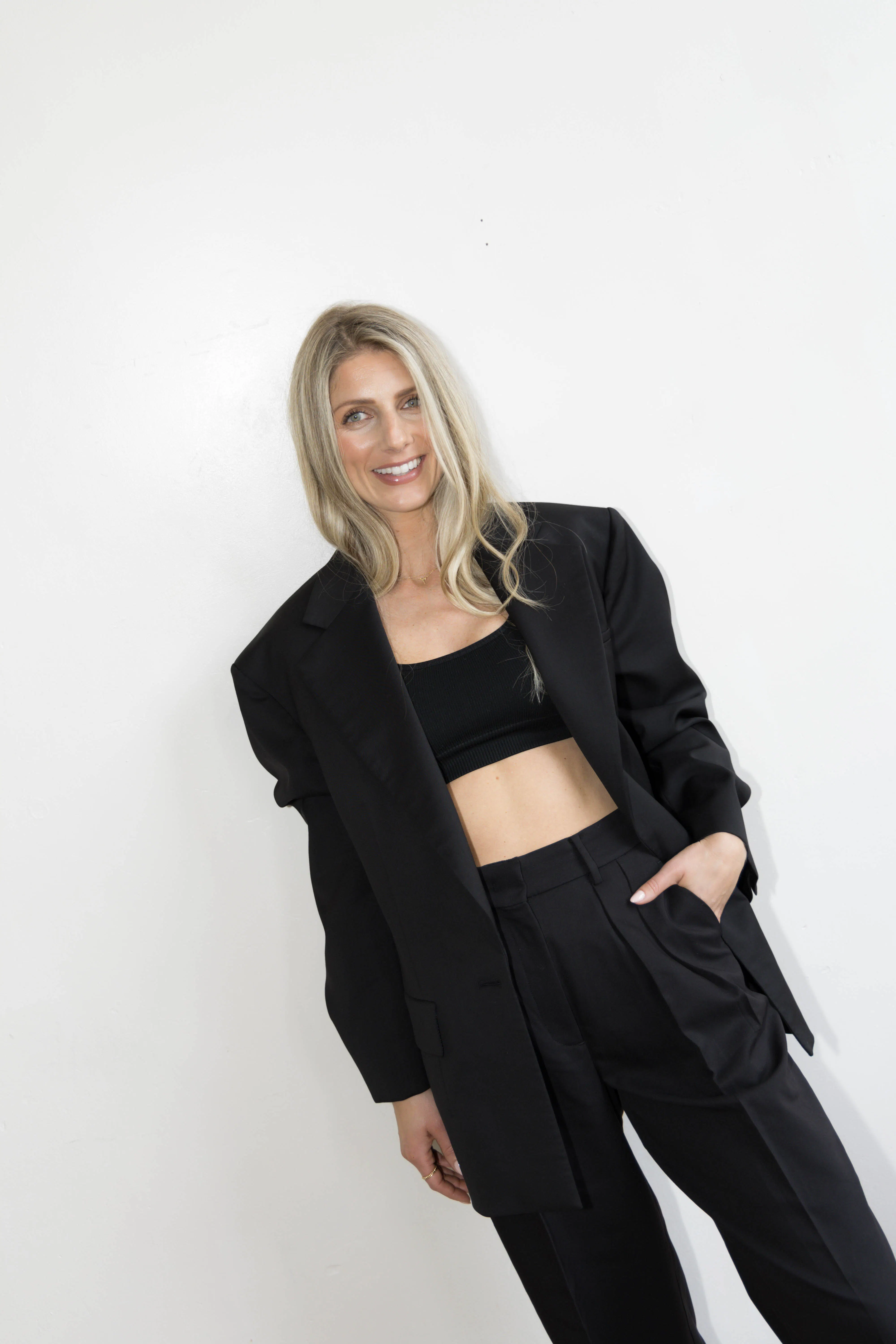 Woman dressed in black trousers, black jacket and black sports bra standing in middle of the picture looking at the camera with hand in pocket on a plain white backdrop.