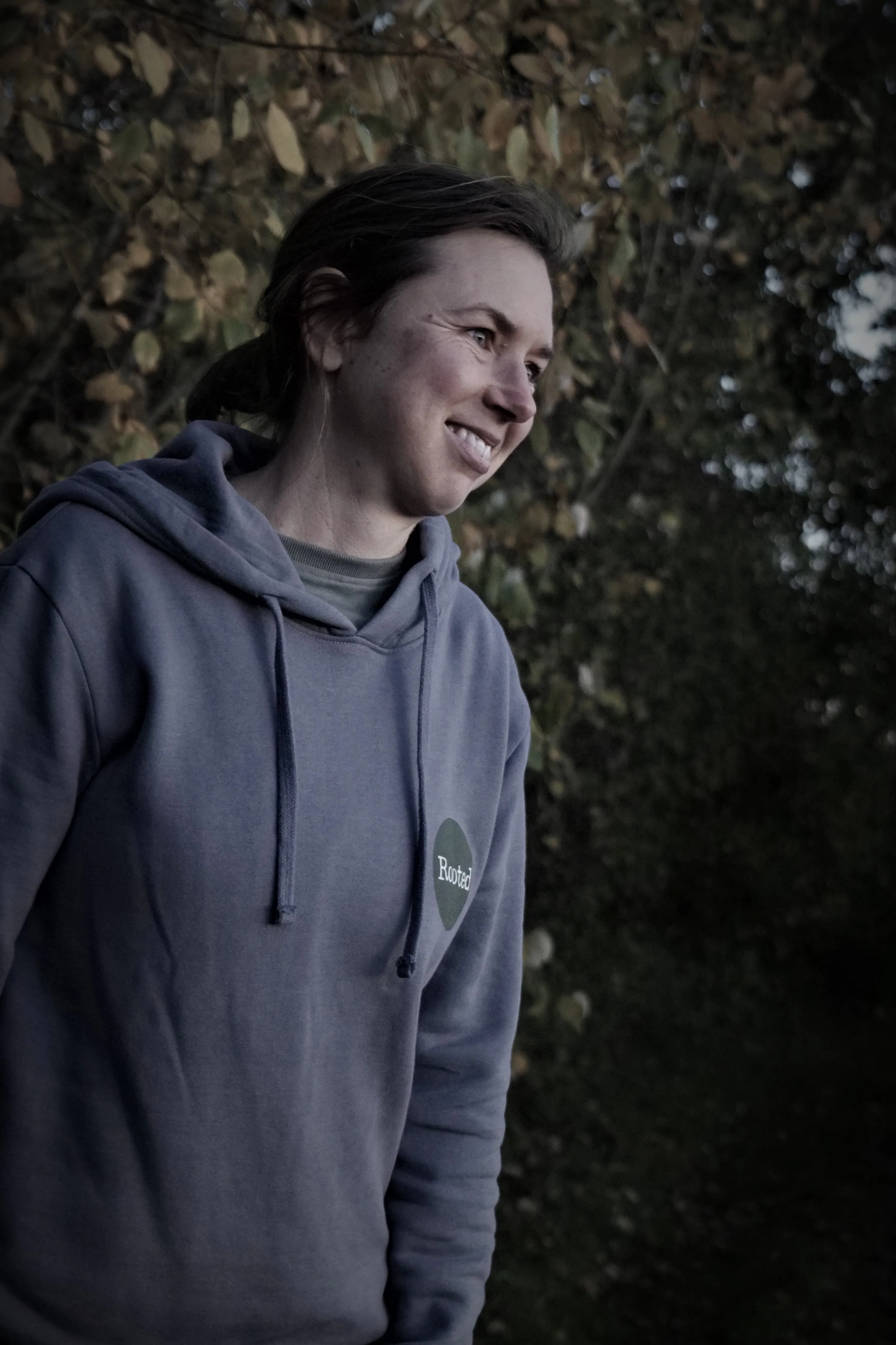 A woman with dark hair is smiling looking away from the camera wearing a hoodie with leaves behind her at twilight. 