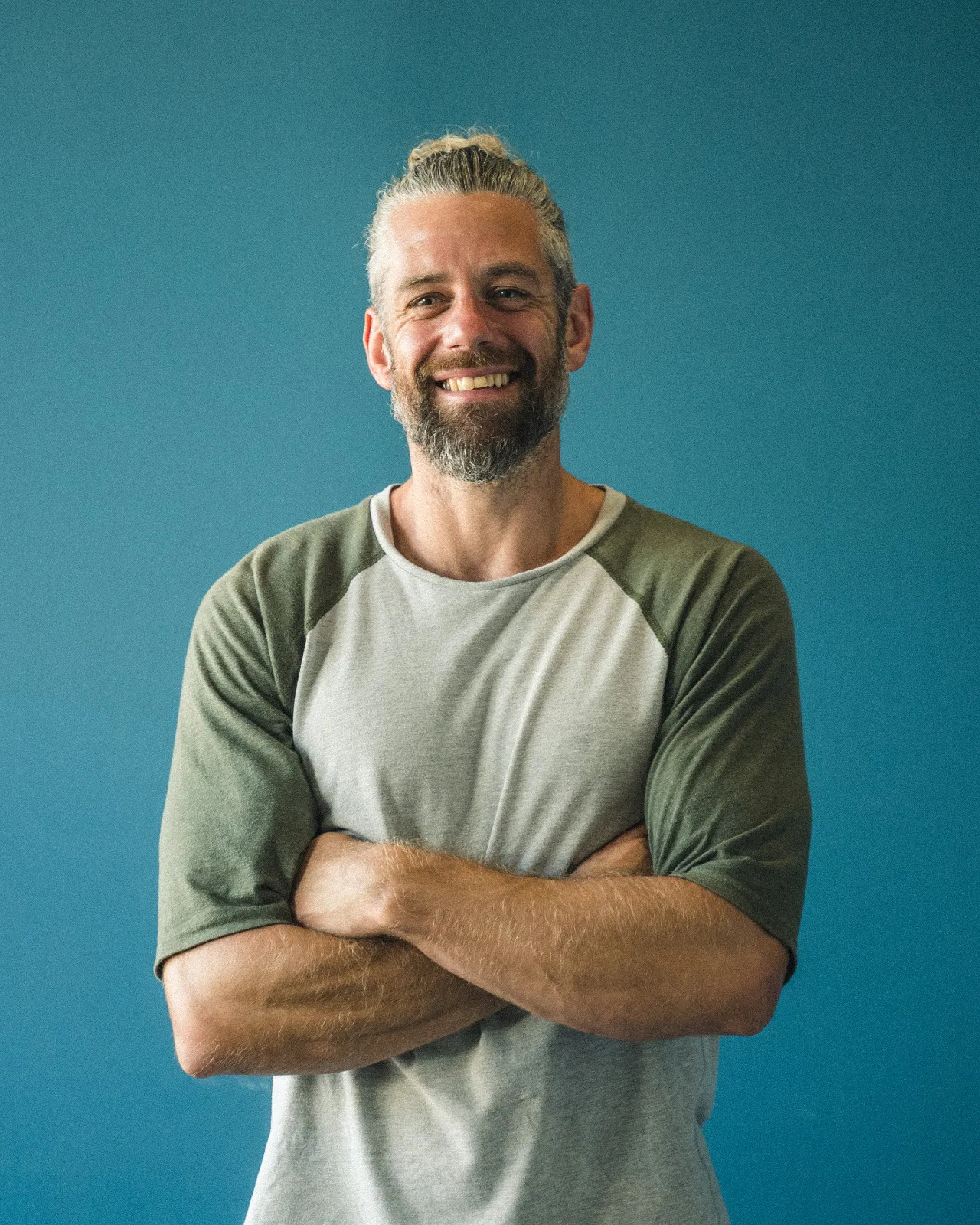 A man with beard is smiling and looking at the camera with his arms folded wearing a loose t-shirt on a plain blue backdrop.