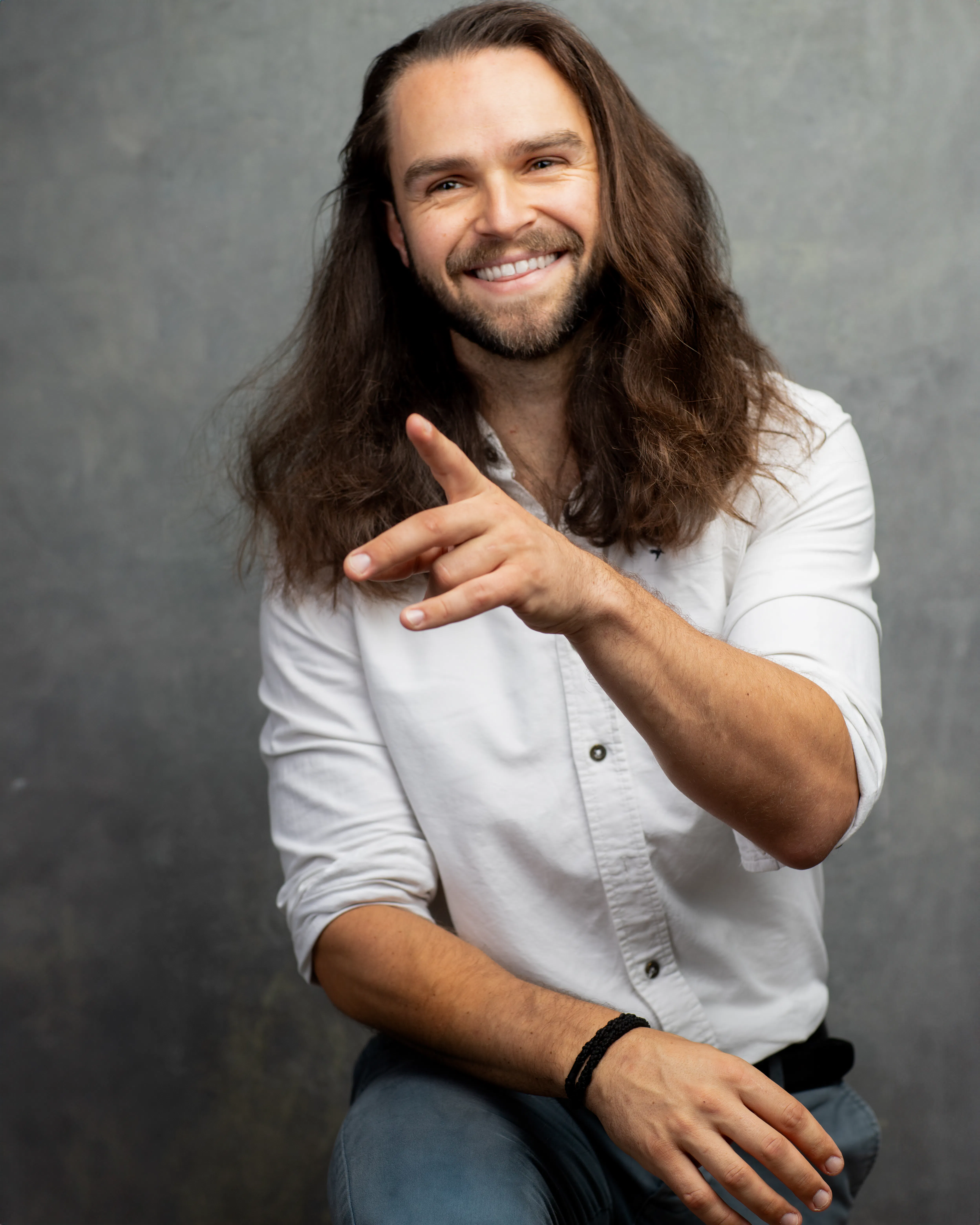 A man with long brown hair, wearing a white shirt, is smiling and pointing towards the camera with his other hand resting on his knee.