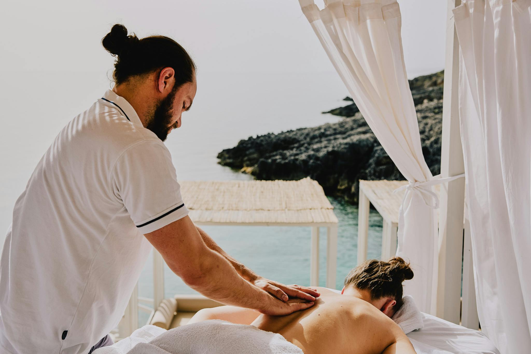 A man in a white polo shirt massages a woman in front of the sea. There are flowing shite curtains and cabanas in the background.