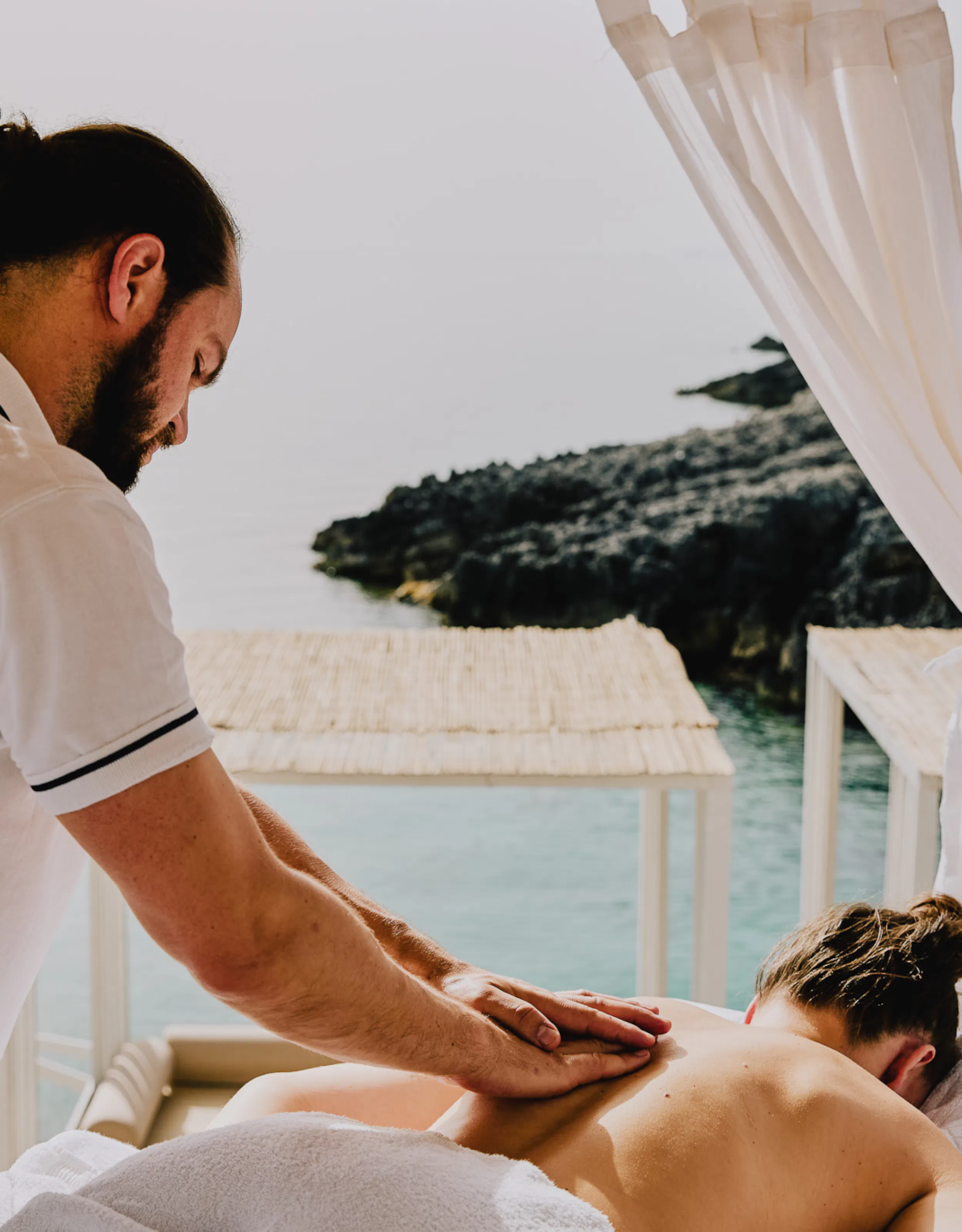 A man in a white polo shirt massages a woman in front of the sea. There are flowing shite curtains and cabanas in the background.