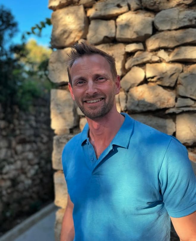 A man in a blue polo shirt smiles into the camera, standing in front of a stone wall with dappled sunlight and bright blue skies in the background.